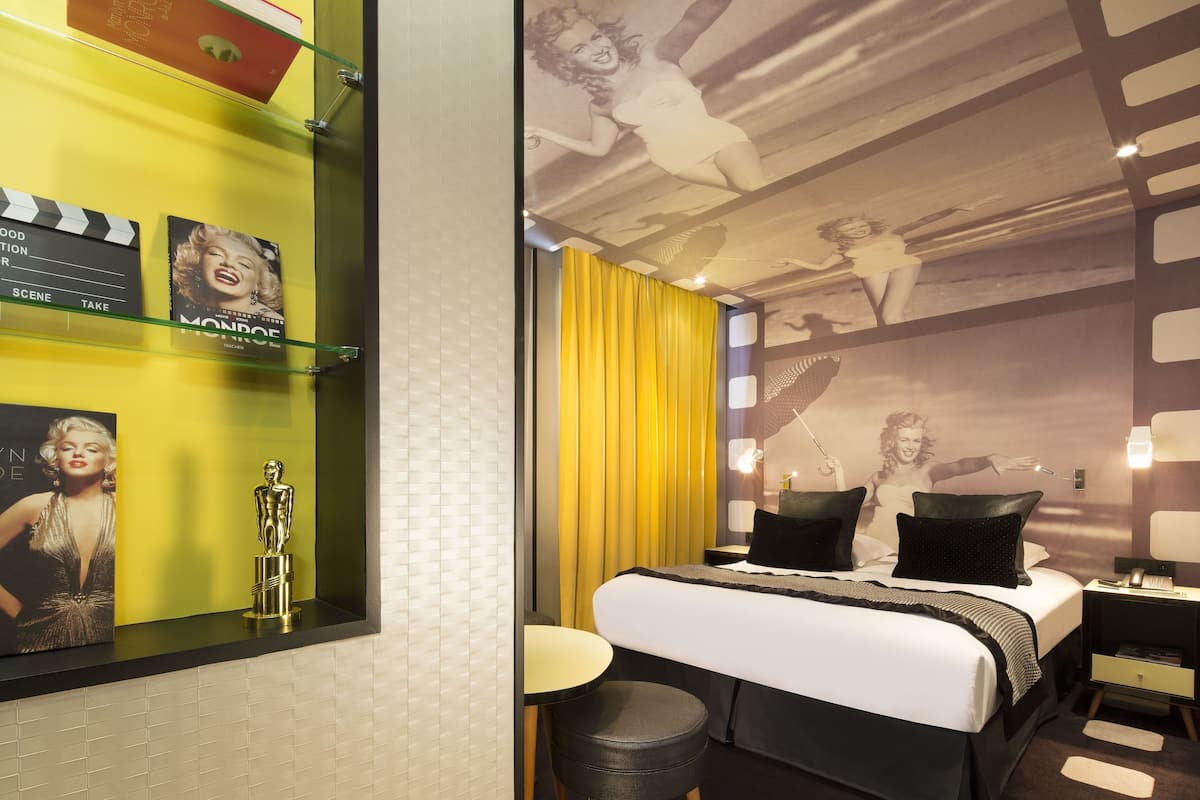 Una habitación de hotel con cama, una mesita, una silla, una mesita de noche y una pared decorada con un póster de cine y una estatua.