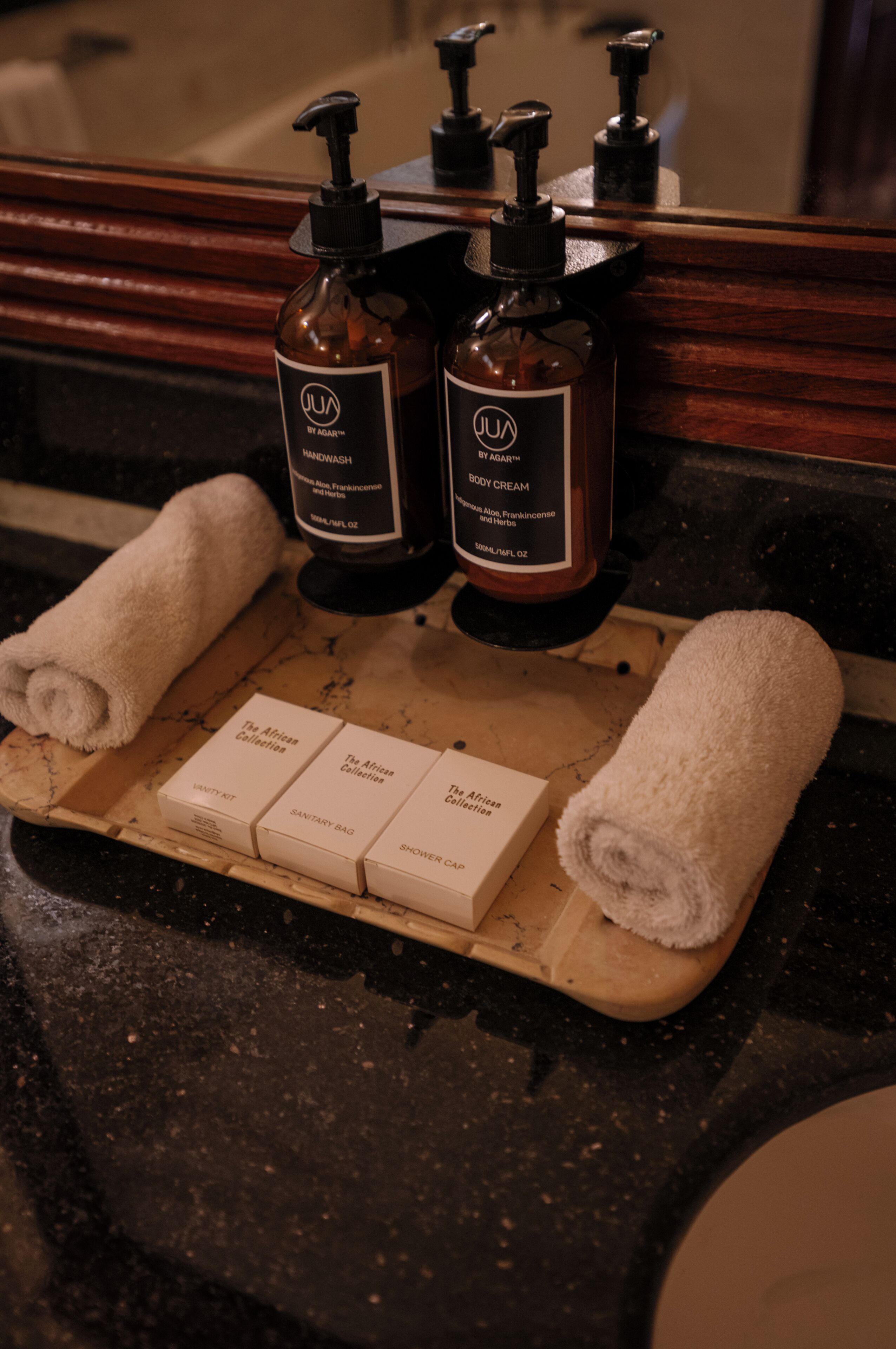 Un comptoir de salle de bain avec deux flacons de crème pour le corps JUA et deux flacons de savon pour les mains JUA, un plateau en bois contenant trois boîtes de produits JUA et deux serviettes enroulées.