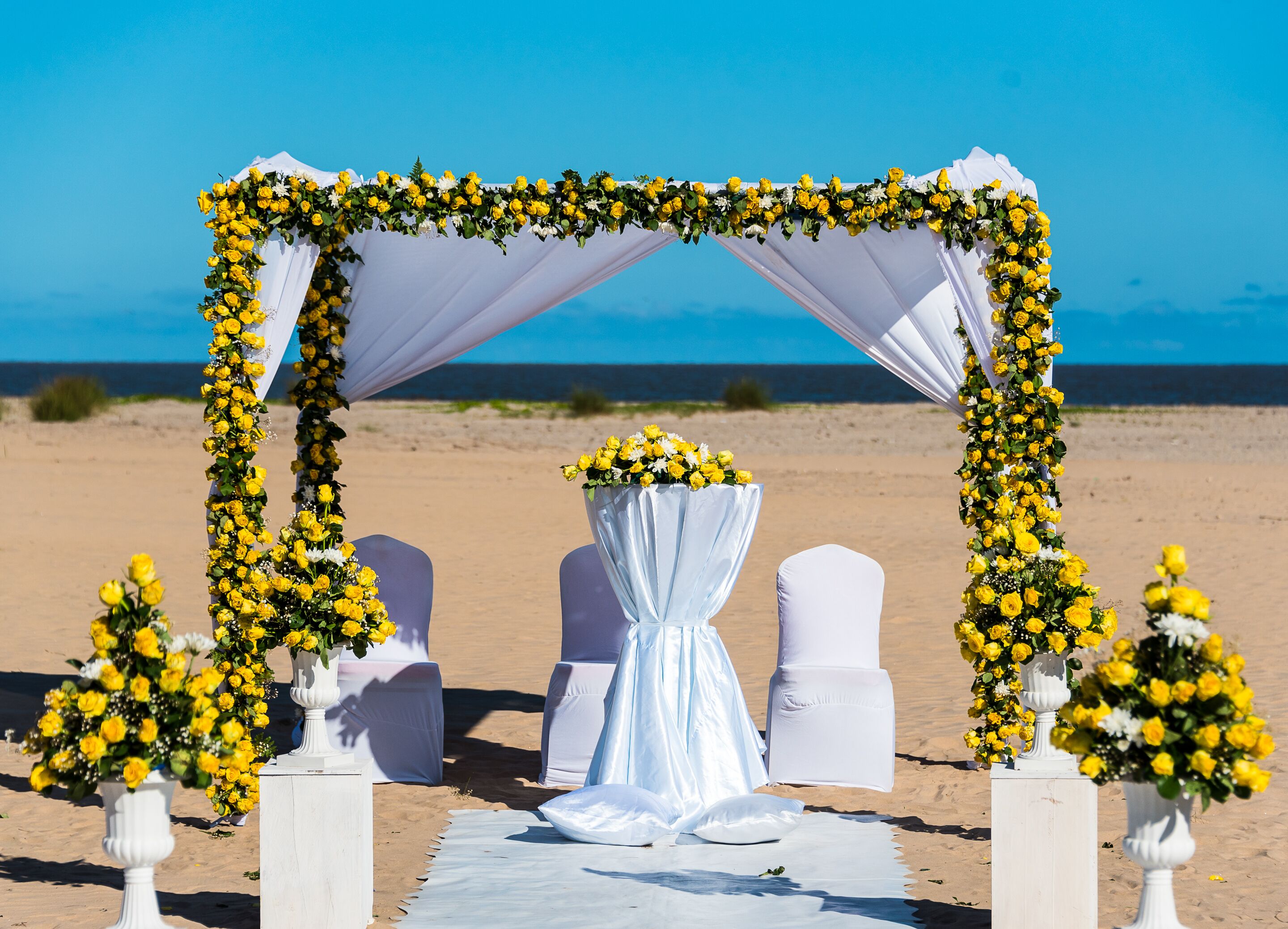 A beach wedding setup with a white arch decorated with yellow flowers, white chairs, and a table with a white tablecloth.