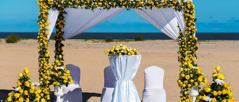 A beach wedding setup with a white arch decorated with yellow flowers, white chairs, and a table with a white tablecloth.