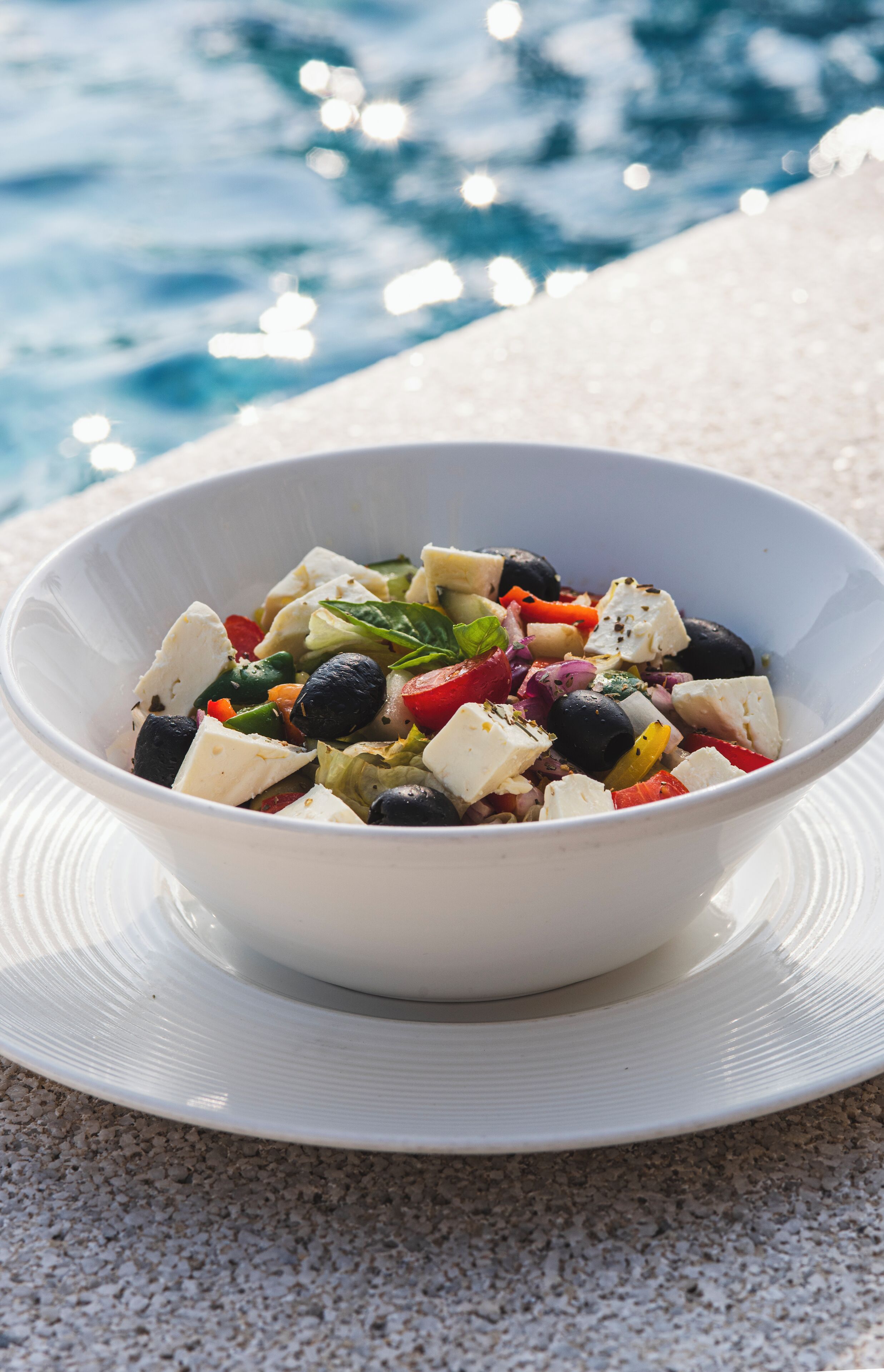 A white bowl of salad with feta cheese, olives, and vegetables on a white plate.