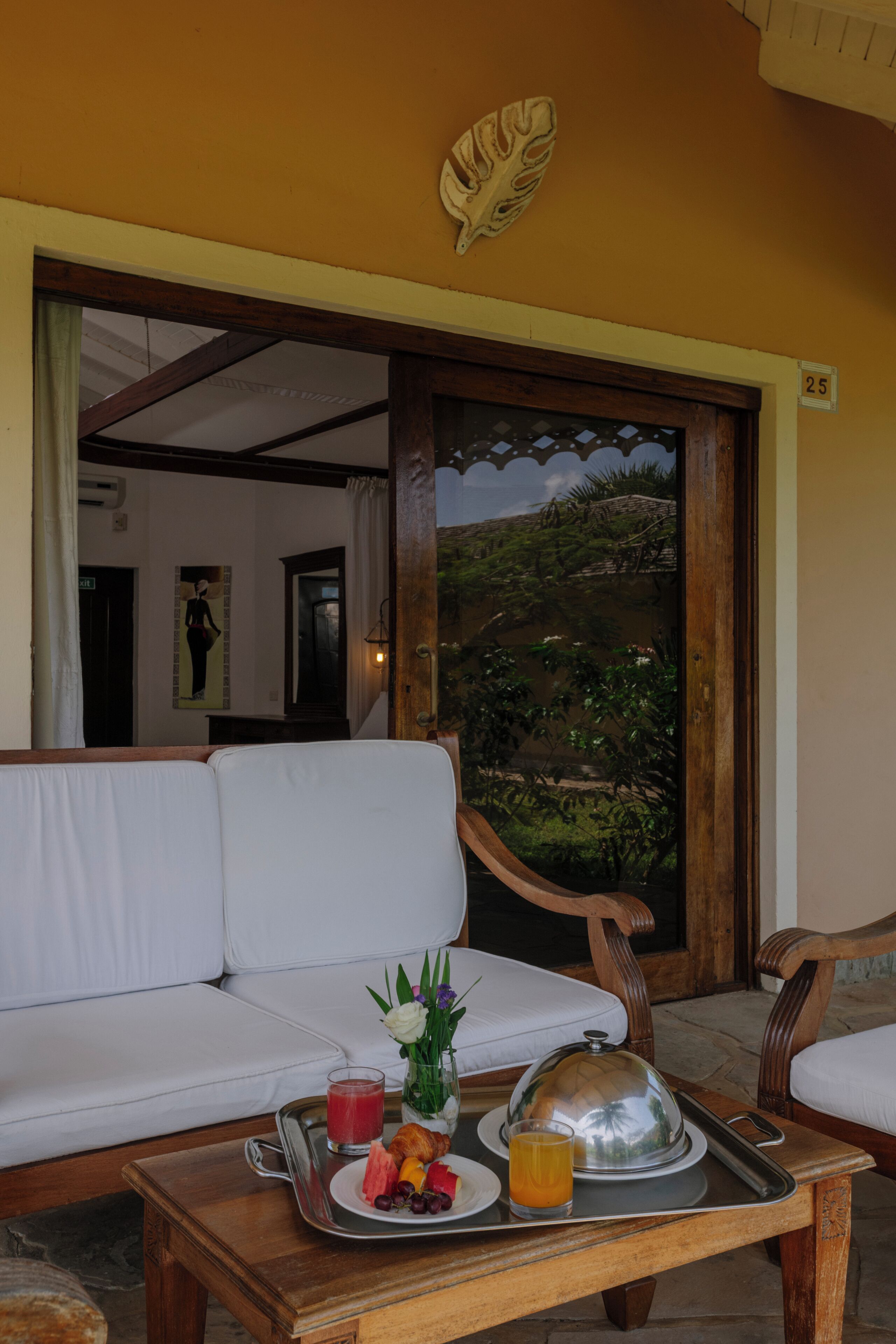Un desayuno servido en una bandeja con comida y bebidas sobre una mesa de madera, un sofá blanco y una puerta de madera con vistas a la vegetación.