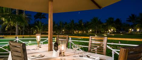 Un área de comedor al aire libre con una mesa dispuesta para dos personas, bajo un toldo, con vista a un jardín ajardinado con palmeras y una vista de la carretera.