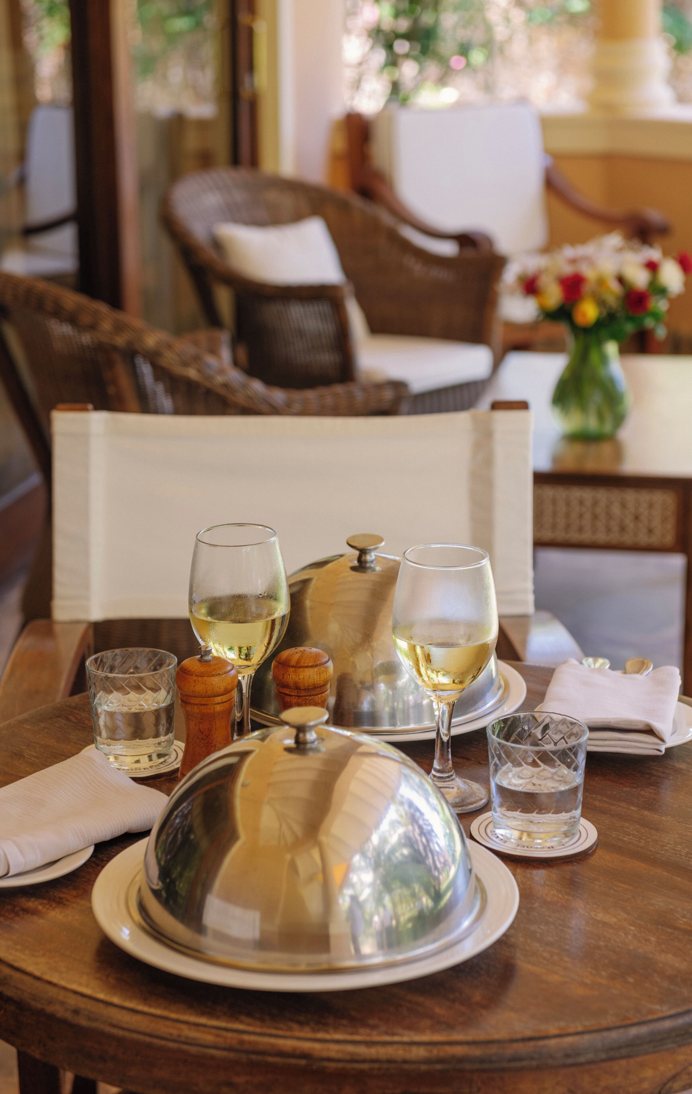A dining table set with wine glasses, a silver cloche, and a small vase with flowers.
