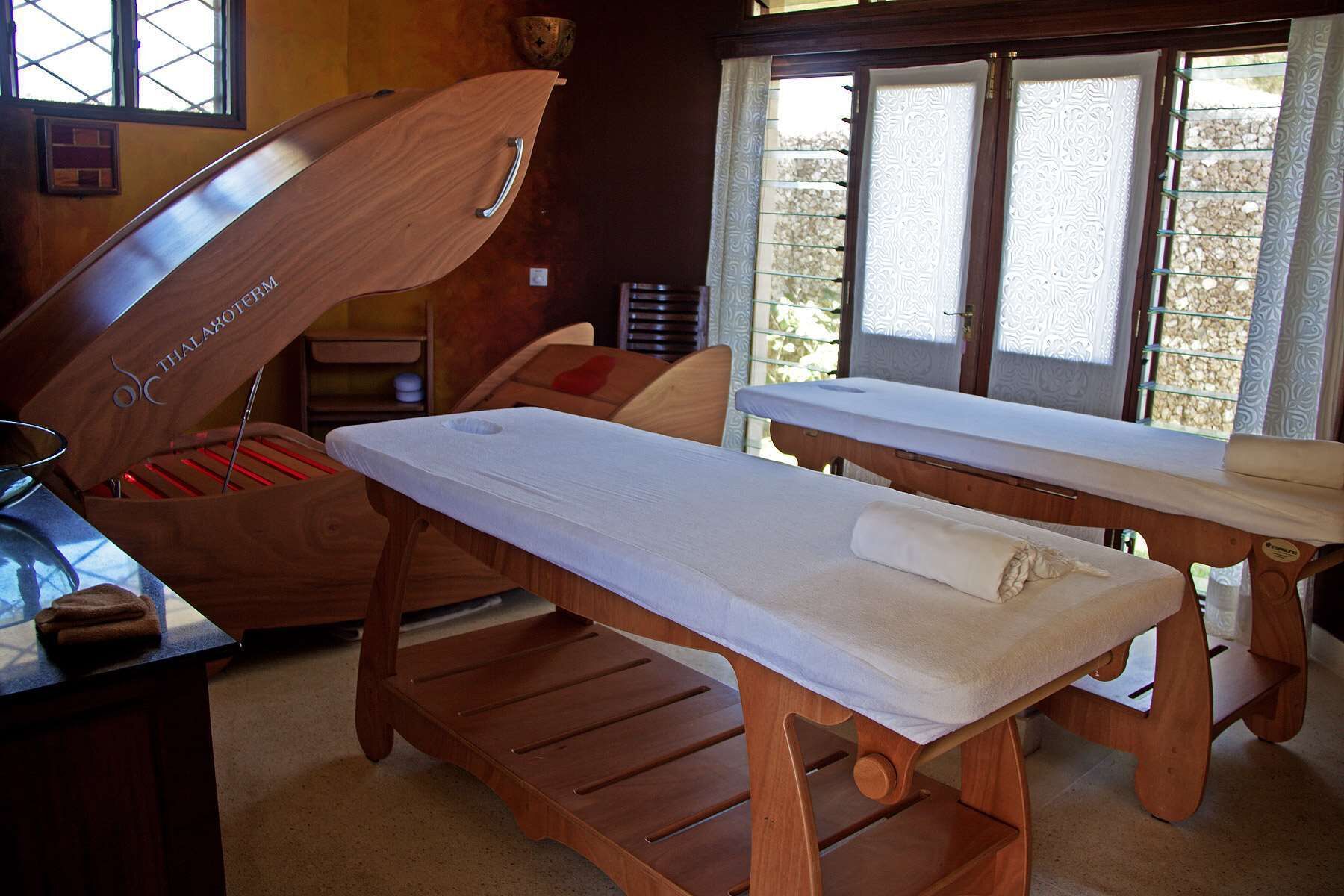 A wooden massage table with a towel, a wooden cabinet with a handle, and a window with patterned curtains.