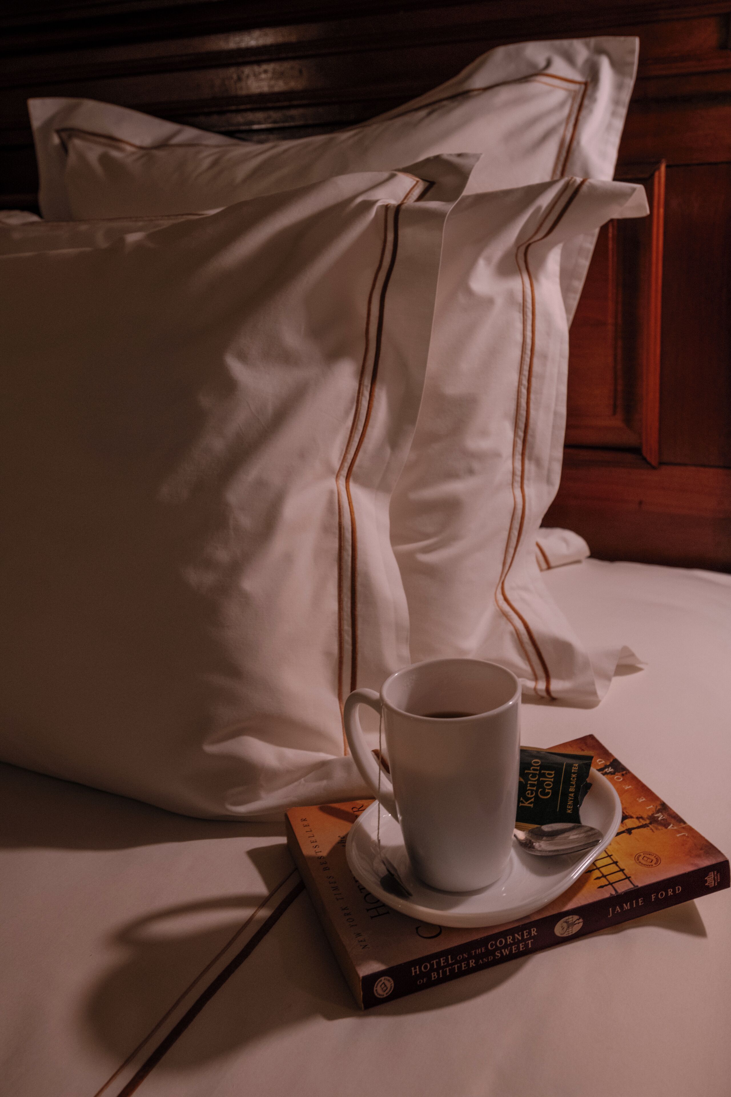 A neatly made bed with white linens and pillows, a cup of coffee on a saucer, and a book titled "Hotel on the Corner of Bitter and Sweet" by Jamie Ford.