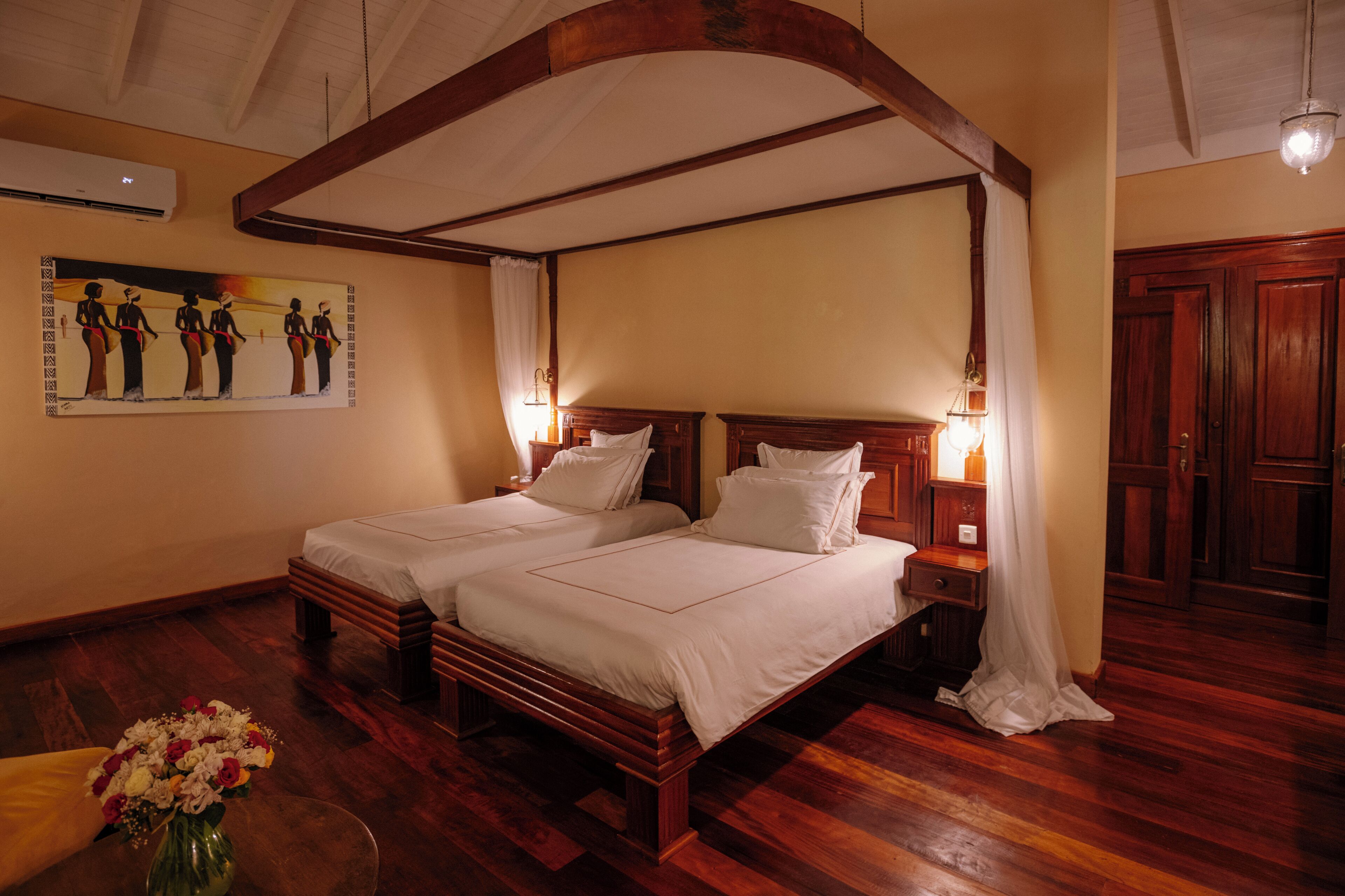 A double bed with a canopy, a bedside table with a lamp, a wall-mounted air conditioner, and a painting on the wall.