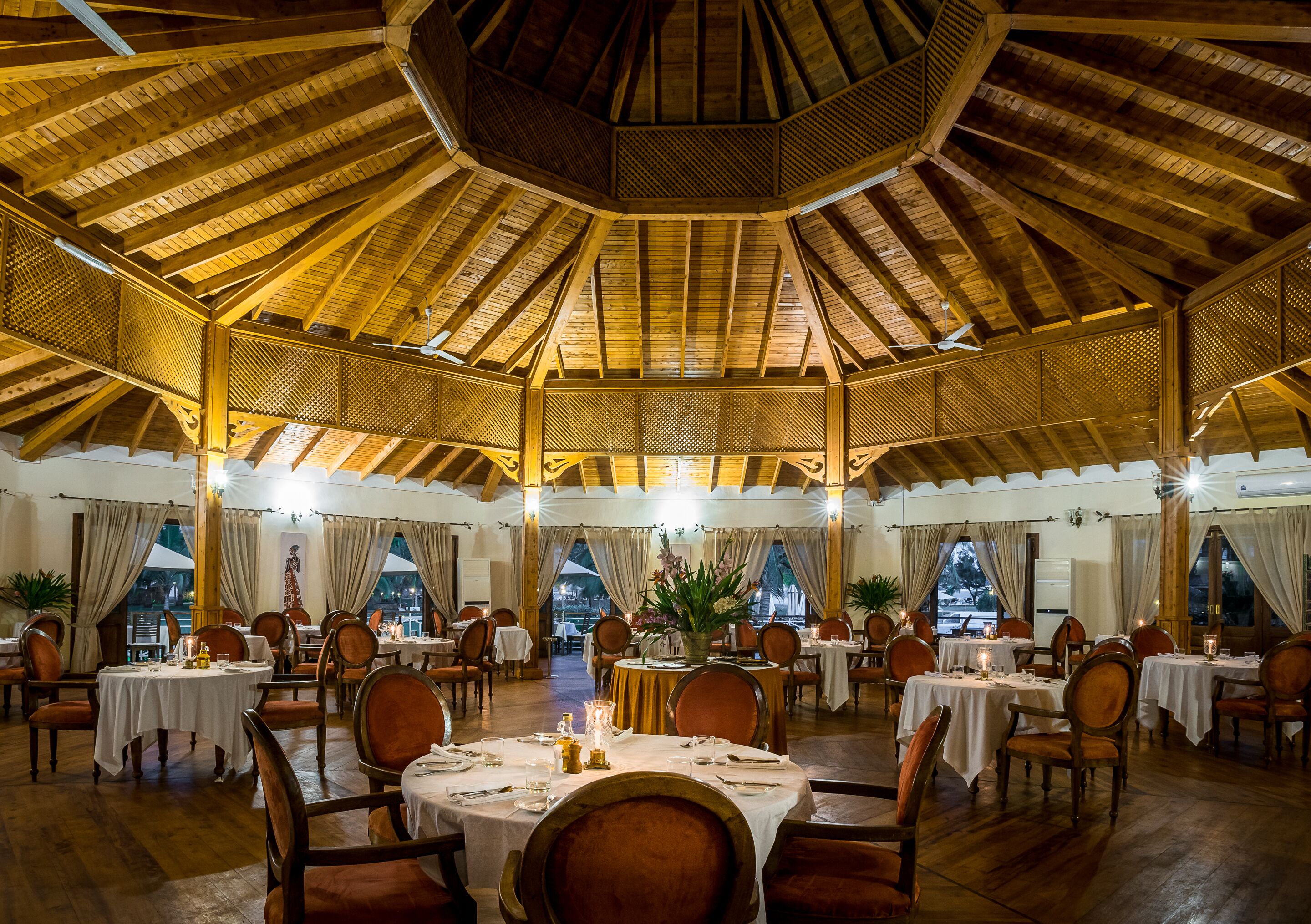 A spacious dining area with multiple tables and chairs, a high wooden ceiling, and large windows.