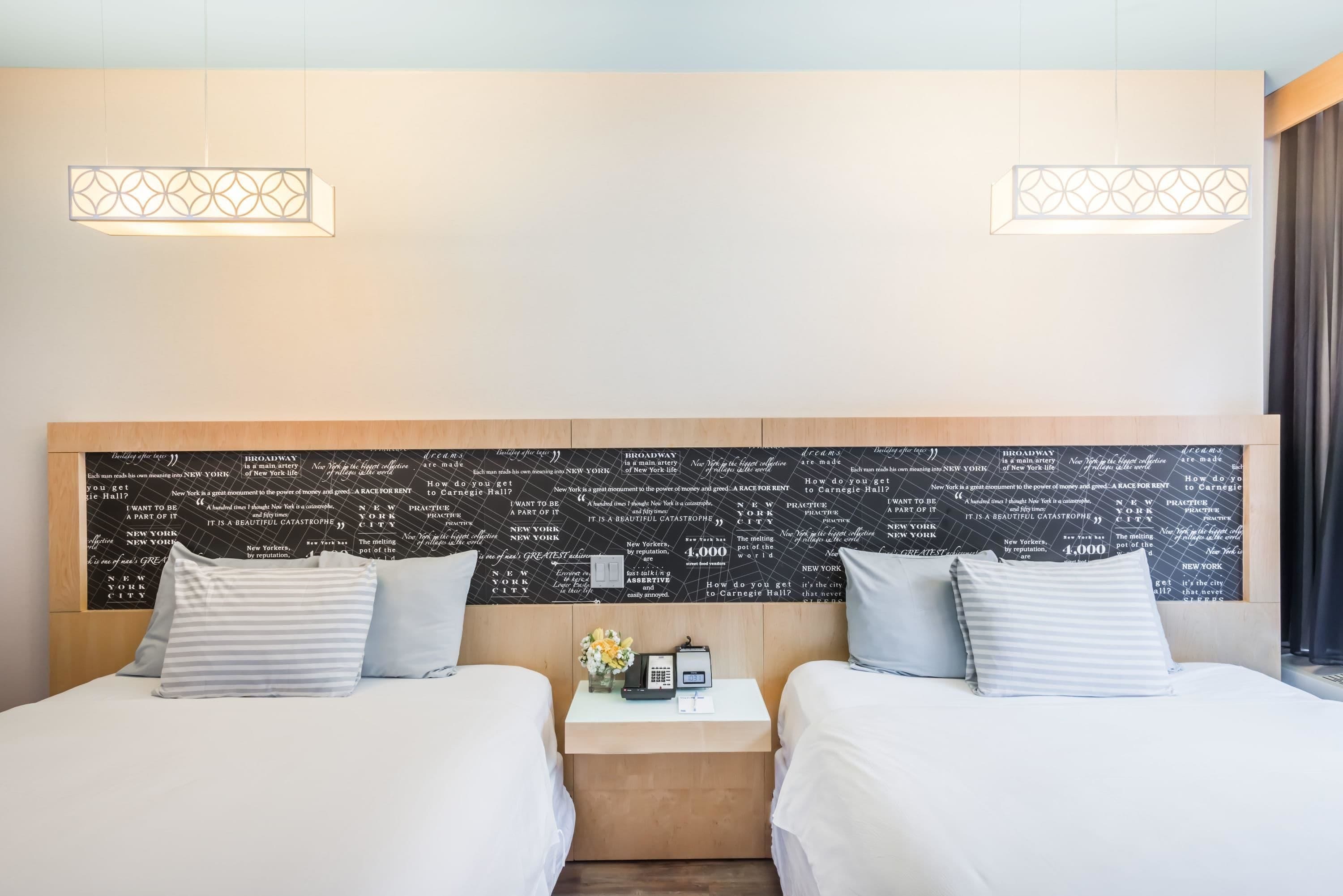 A hotel room with two beds, a bedside table with a phone and a vase of flowers, and a wall with text.