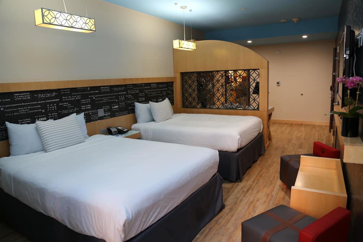 Ein Hotelzimmer mit zwei Betten, einem hölzernen Kopfende mit dekorativer Platte, einer kleinen Sitzecke mit Tisch und einem an der Wand montierten Fernseher.