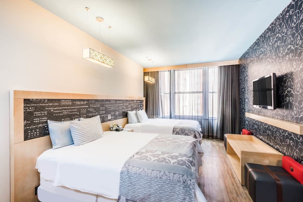 Ein Hotelzimmer mit zwei Betten, einem an der Wand befestigten Fernseher und einem Fenster mit Vorhängen.