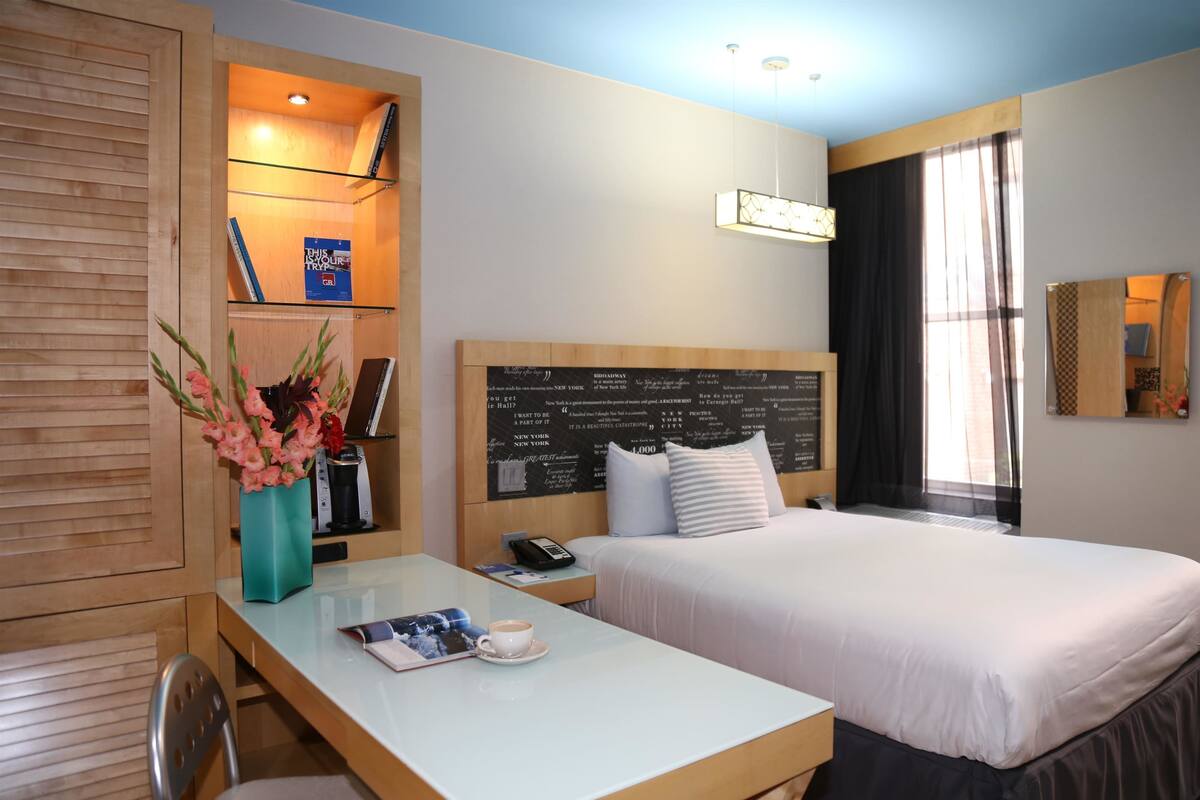 Ein Hotelzimmer mit Bett, Schreibtisch mit Stuhl, Nachttisch, Blumen in einer Vase, Lampe und Fenster mit Vorhängen.