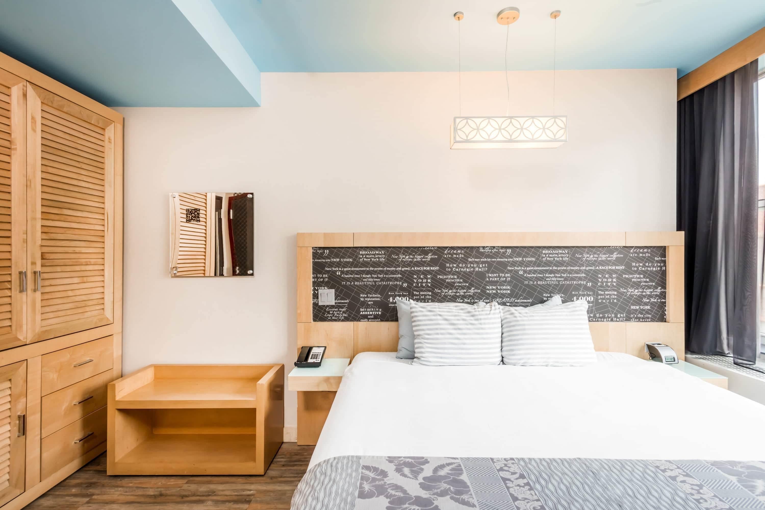 Habitación de hotel con una cama grande, armario de madera, mesita de noche y una pared con una pizarra temática de Broadway.