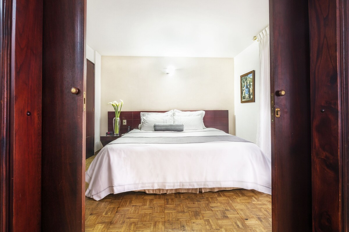 A hotel room with a wooden floor, a bed with white bedding, a nightstand with a vase of flowers, and a painting on the wall.