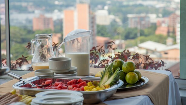 Una mesa de desayuno con frutas, leche y una jarrón, con vista a la ciudad.