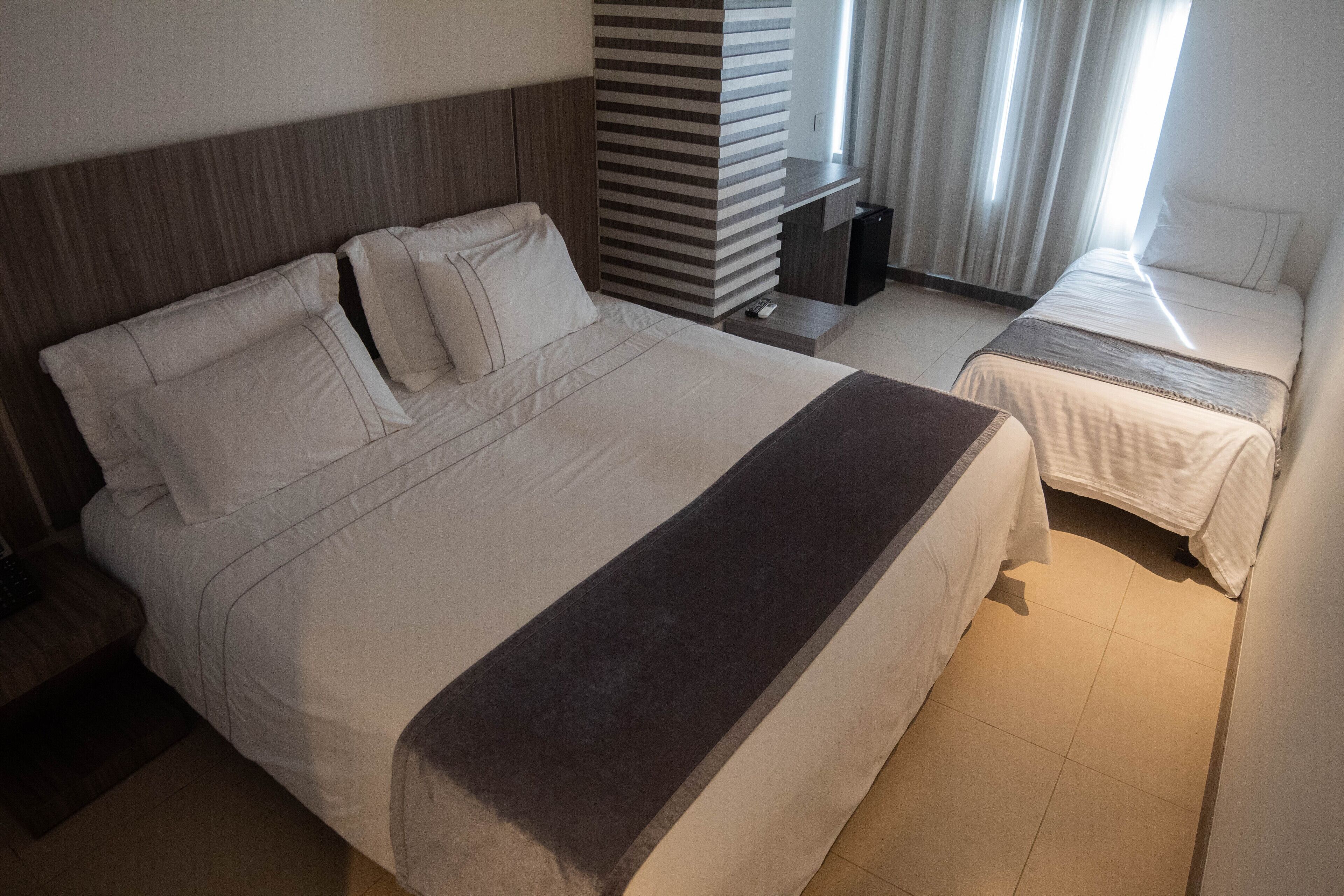 Habitación de hotel con una cama grande, una cama más pequeña, una mesita de noche, un televisor y una ventana con cortinas.