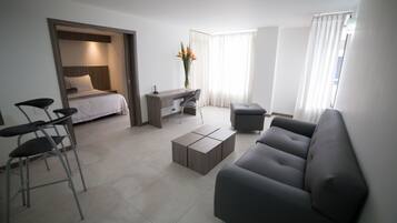 A modern, well-lit living room with a grey sofa, a coffee table, a desk with a chair, and a bed with a headboard.
