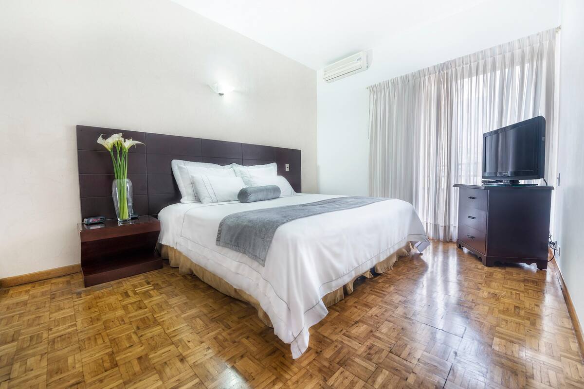 Habitación de hotel con suelo de parquet de madera, cama con ropa blanca, cabecero oscuro, mesita de noche con un jarrón de flores, televisor sobre un mueble y una ventana con cortinas.