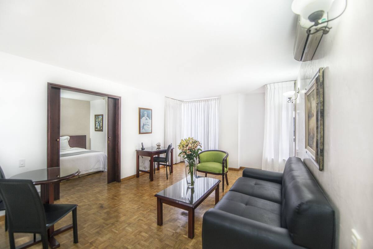 Habitación de hotel con una cama, una mesa de comedor, un sillón verde, un sofá negro y un jarrón con flores.