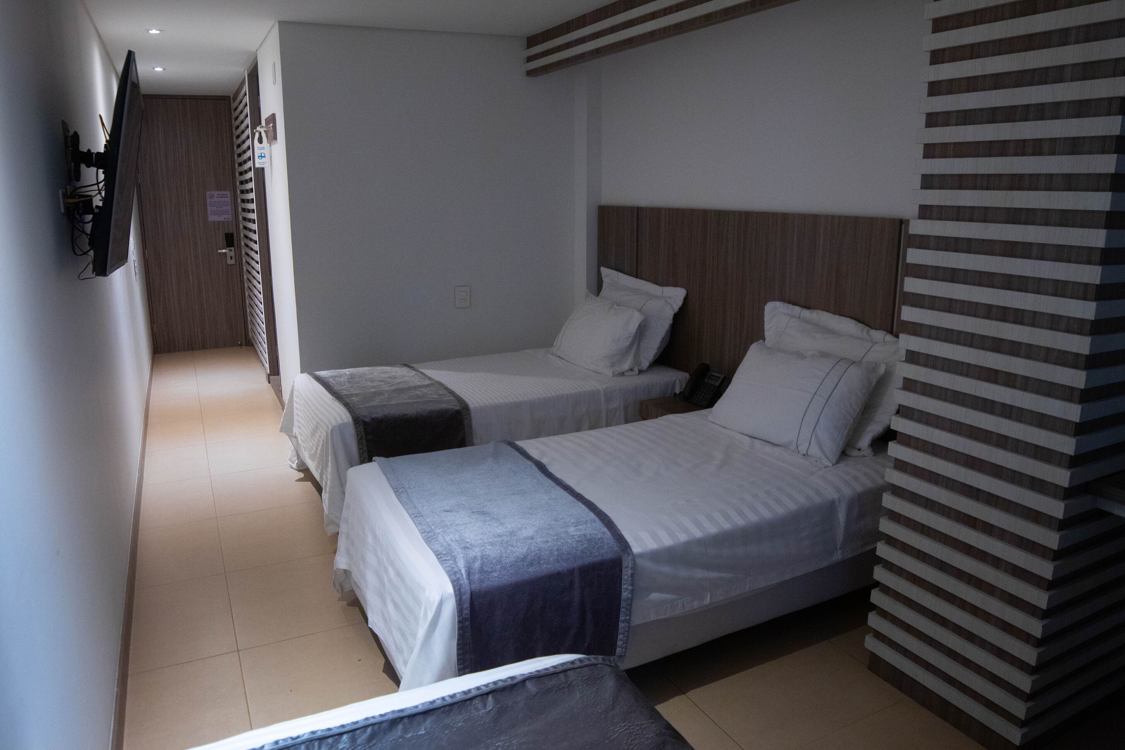 Une chambre d’hôtel avec deux lits simples, une tête de lit en bois, un téléviseur fixé au mur et une porte donnant sur un couloir.