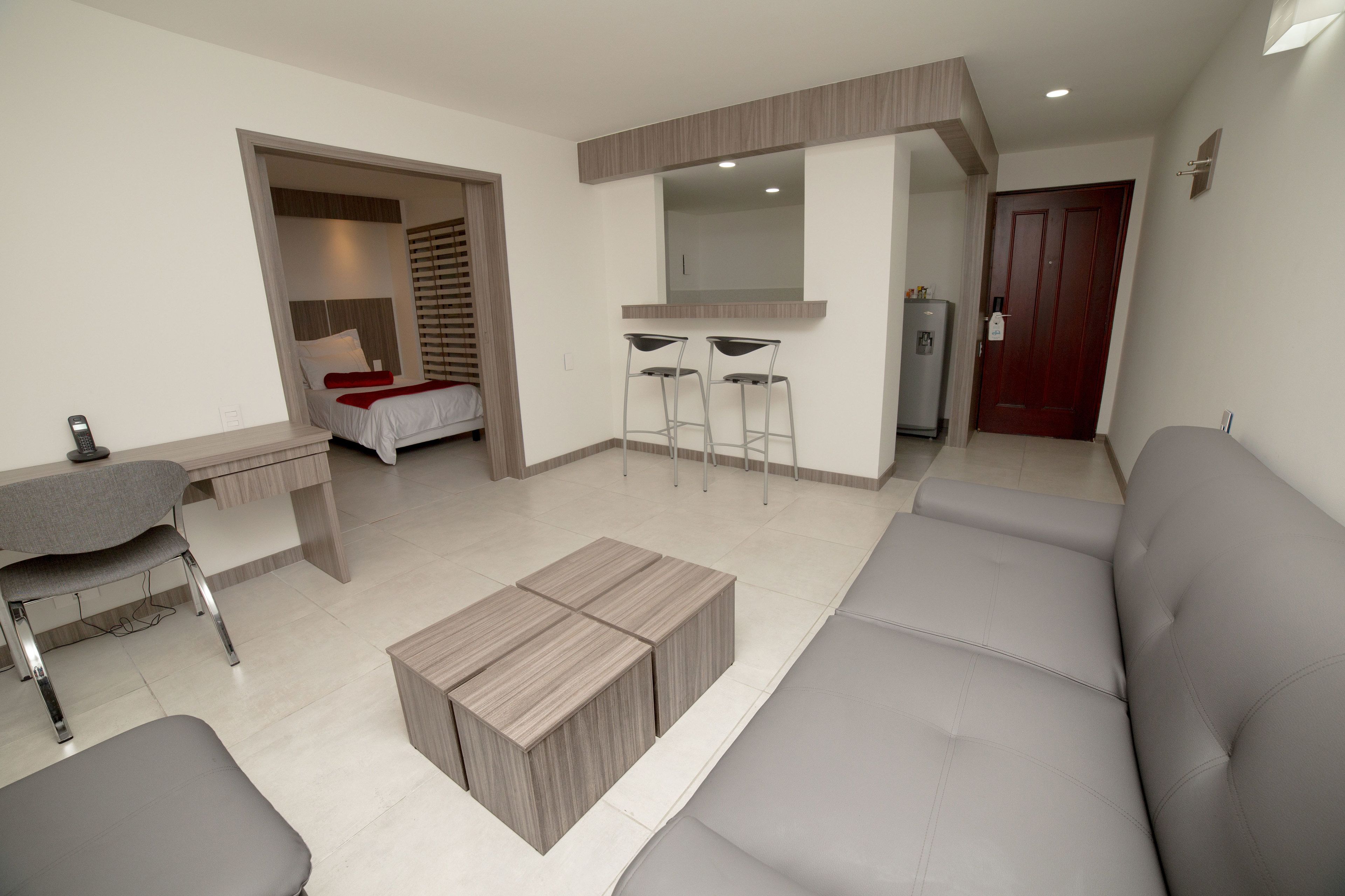 Un salon moderne avec un canapé gris, une table basse en bois et un bureau avec une chaise. On y trouve également une petite kitchenette équipée d’un réfrigérateur et dotée d’une porte en bois.