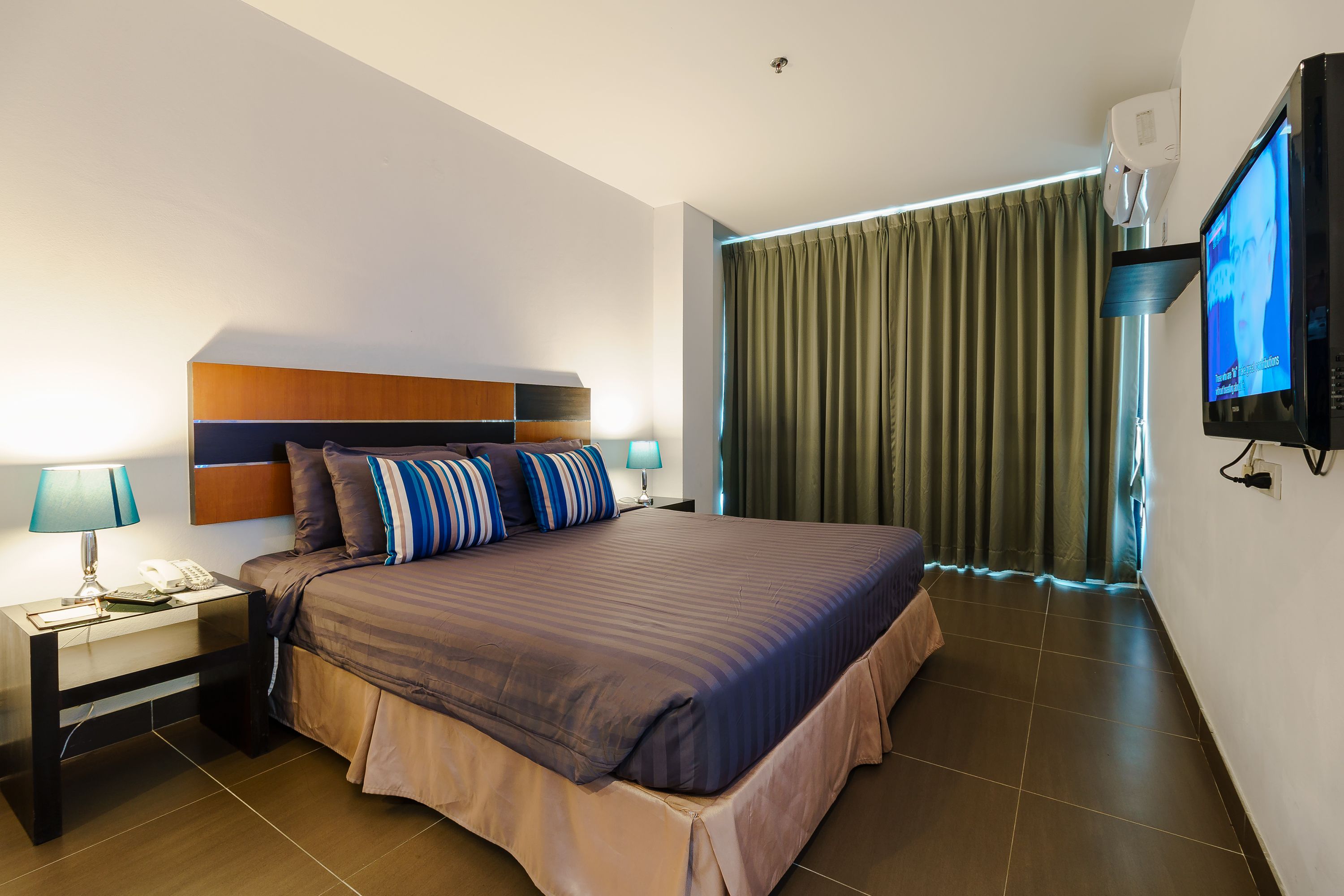 Une chambre d’hôtel avec un grand lit, une table de chevet avec une lampe, un téléviseur fixé au mur et une fenêtre avec des rideaux.