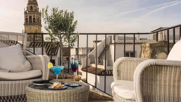 A rooftop terrace with wicker furniture, a table with snacks and drinks, and a view of a historic tower.