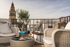Terraza en la azotea con mobiliario de mimbre, una mesa con aperitivos y bebidas, y vistas a una torre histórica.