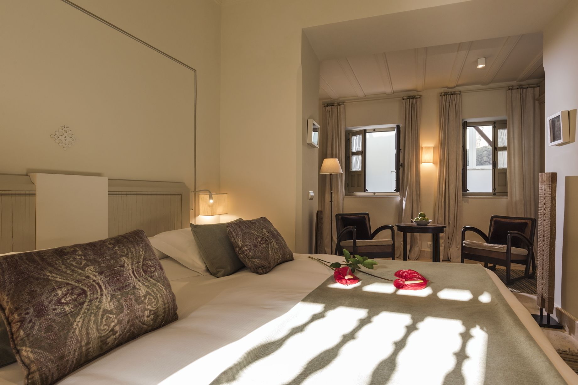 Habitación de hotel con una cama, lámparas de noche, una mesita con un frutero y una planta, y dos sillas.