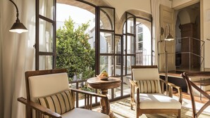 A room with two wooden chairs with striped cushions, a small round table with a bowl of fruit, and a large window with a view of greenery outside.