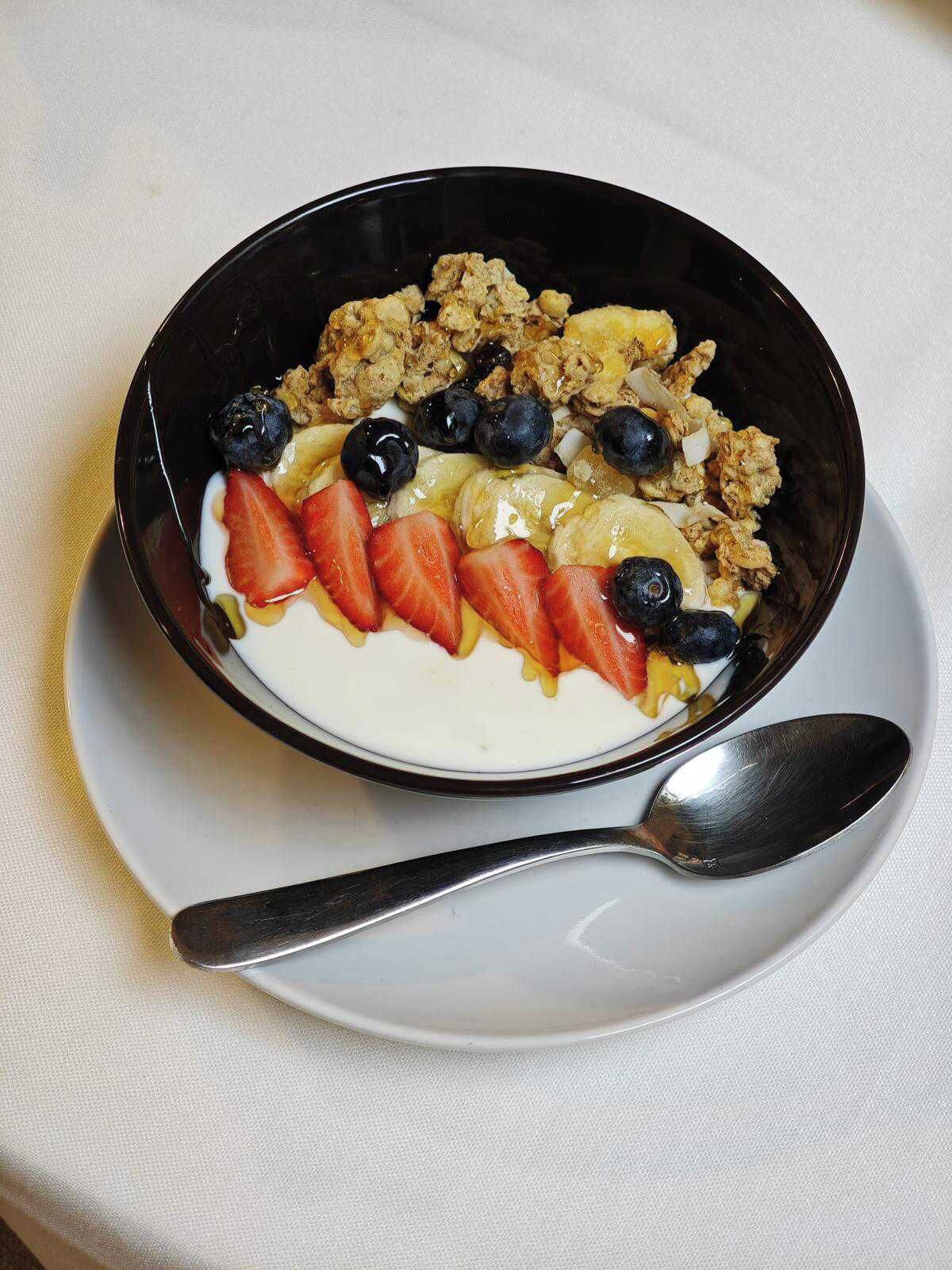 A bowl of yogurt topped with granola, strawberries, blueberries, and banana slices on a white plate with a spoon.