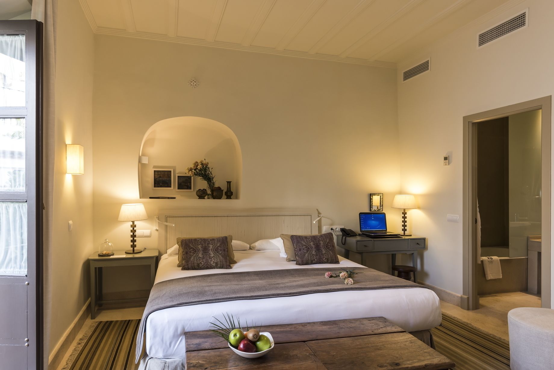 Une chambre d’hôtel avec un grand lit, deux tables de chevet avec des lampes, un petit bureau avec un ordinateur portable, un miroir et un panier de fruits sur une table en bois.