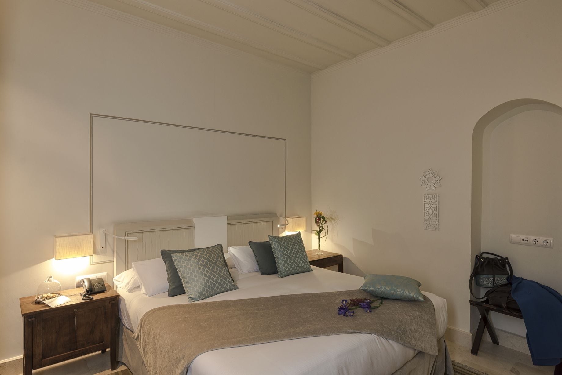 A hotel room with a bed, bedside tables, a lamp, a vase with flowers, and a wall-mounted light fixture.