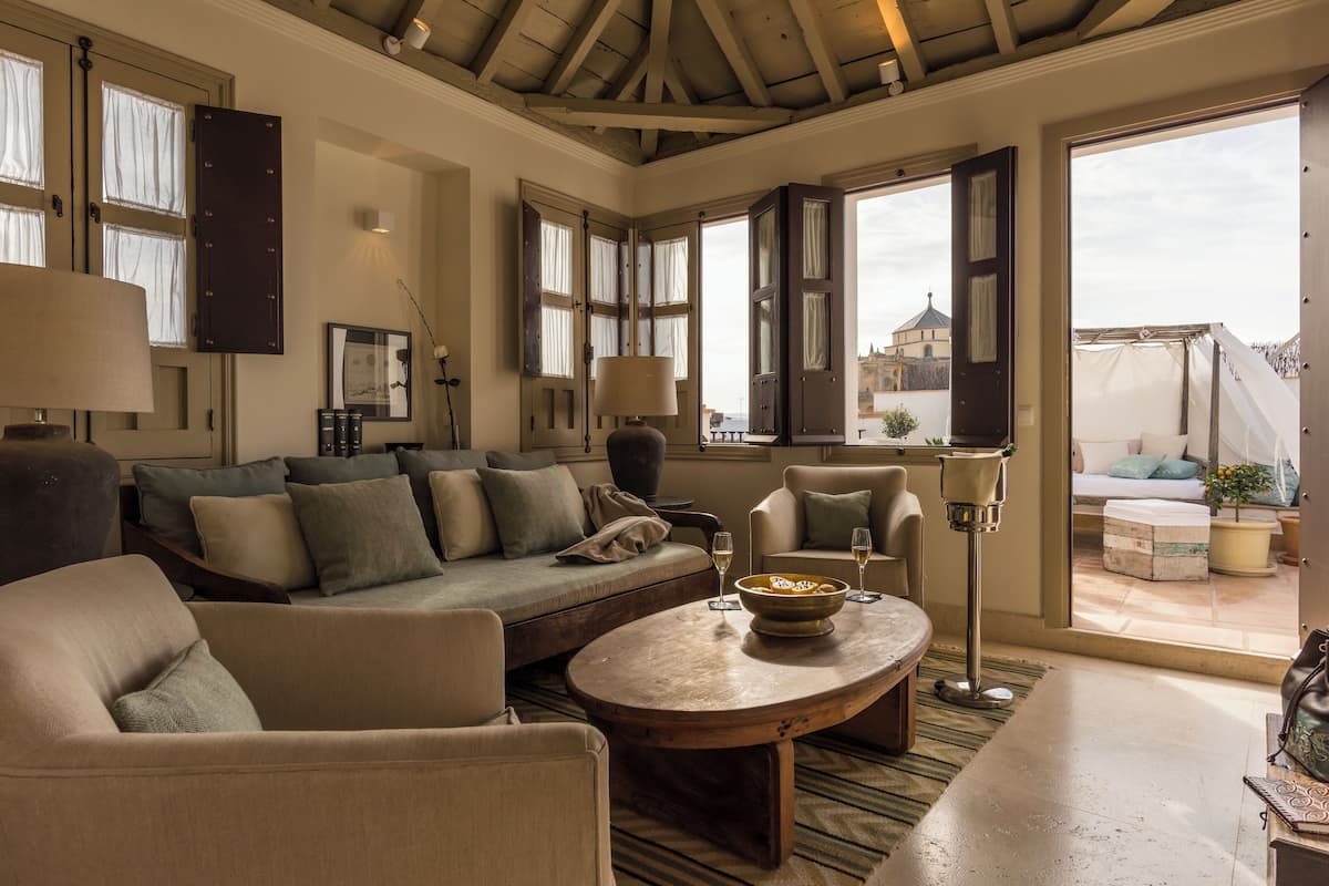 A cozy living room with a sofa, armchairs, a coffee table, and a view of the city through open doors.