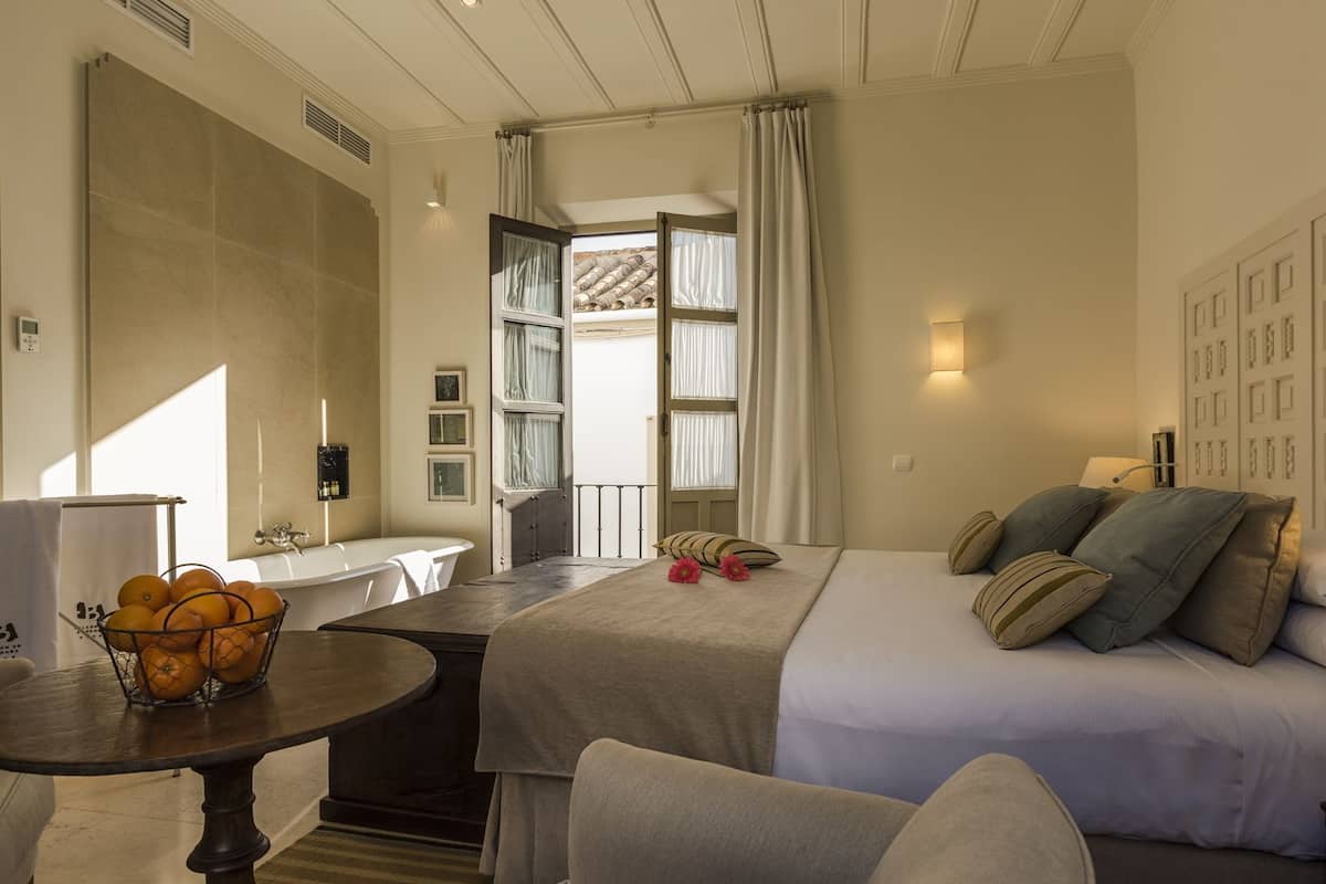 A hotel room with a bed, a small table with oranges, a sink, and a window with curtains.