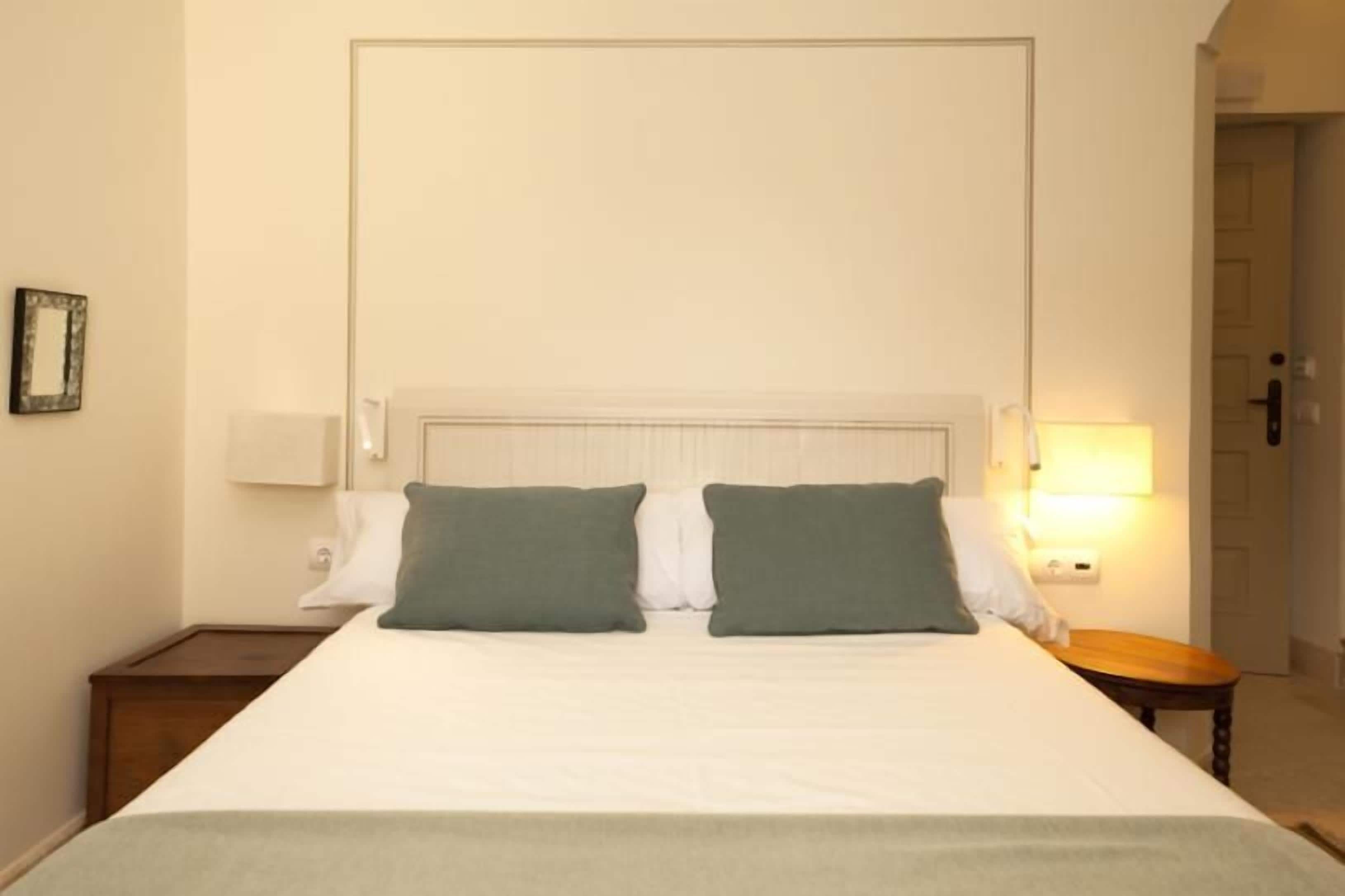 A neatly made bed with white linens and two green pillows, a bedside table with a lamp, and a framed picture on the wall.