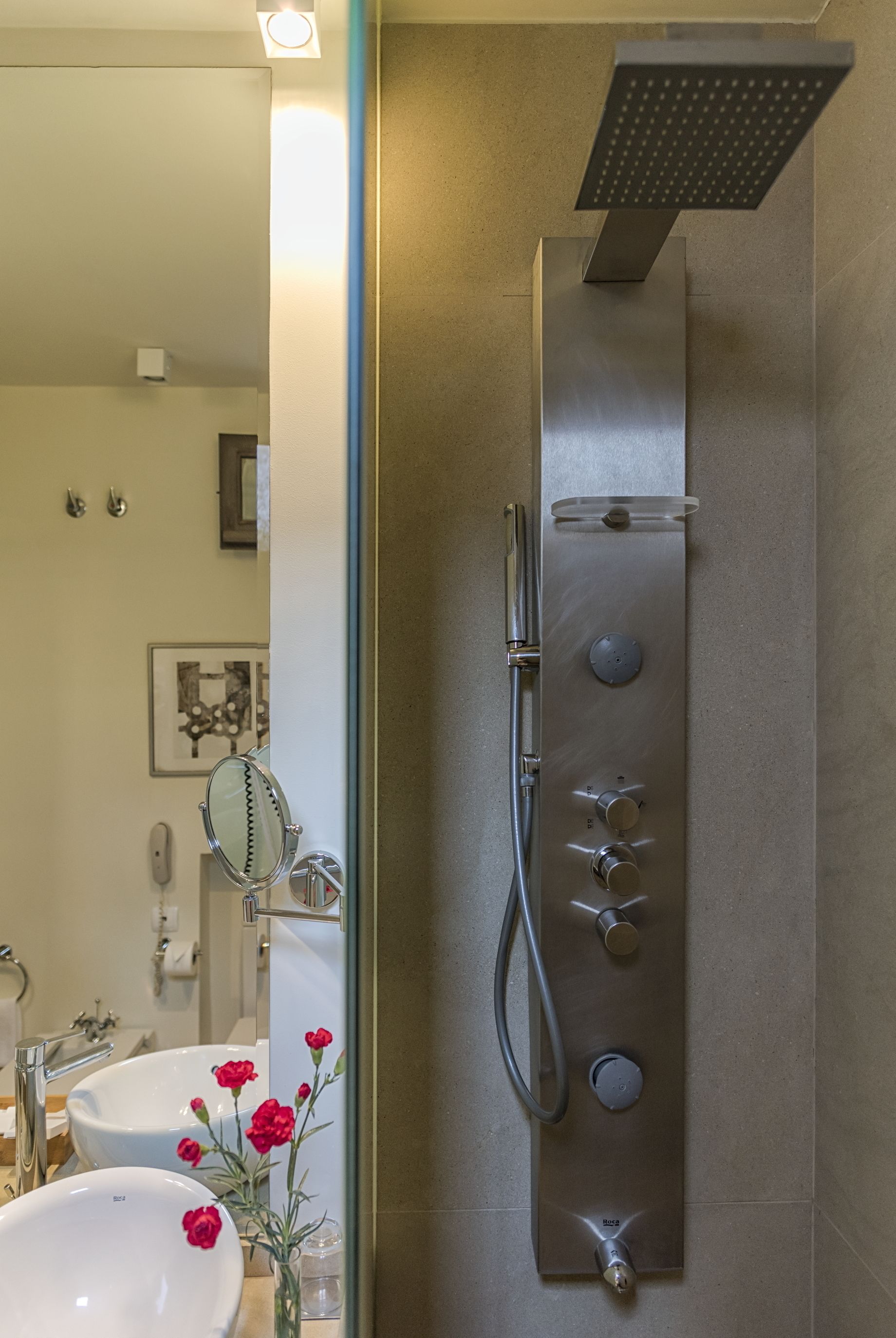 A modern bathroom with a large mirror, two sinks, a shower with multiple controls, and a vase with red flowers.