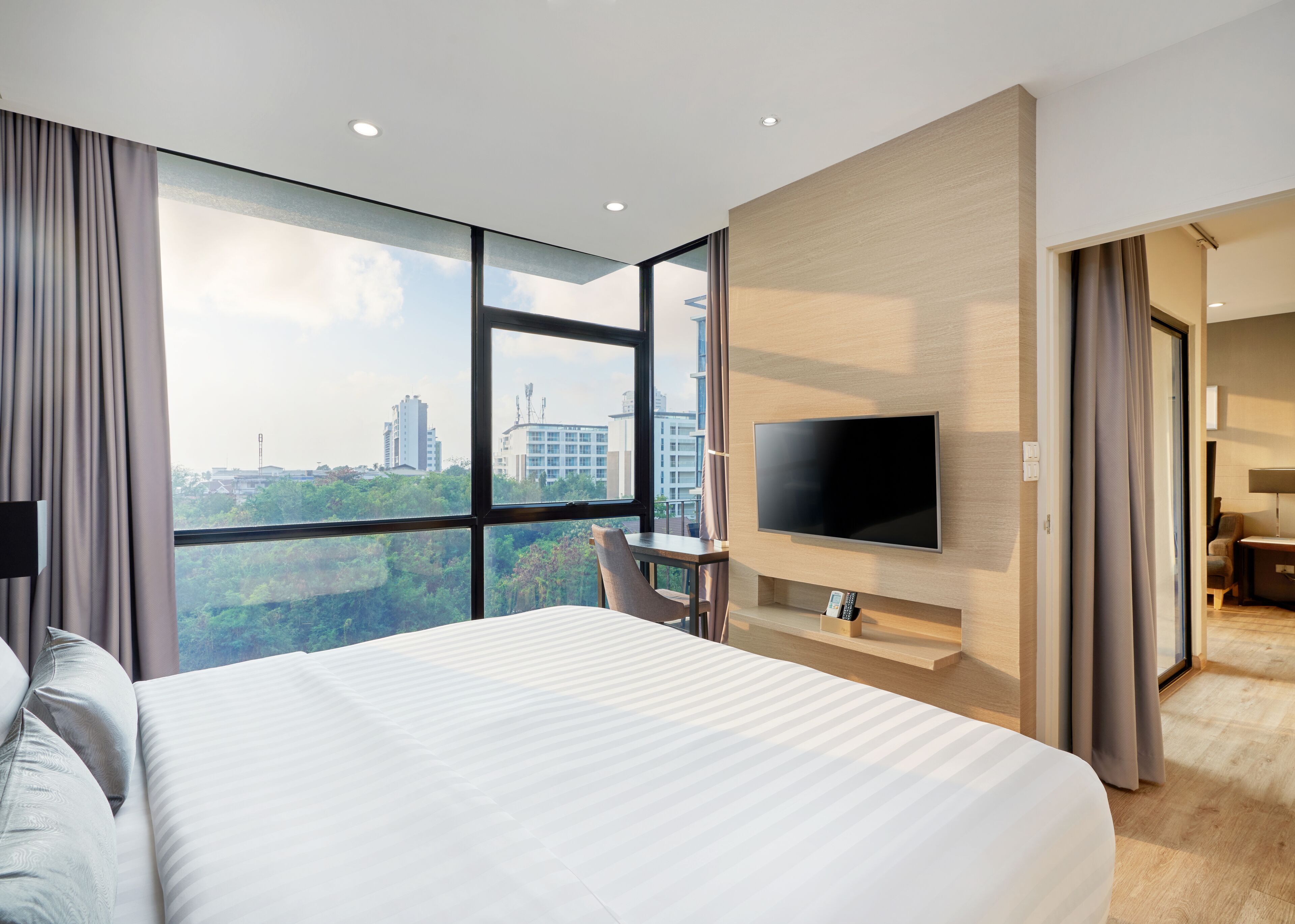 Una habitación de hotel moderna con una cama grande, un televisor montado en la pared y vistas a la ciudad a través de amplios ventanales.