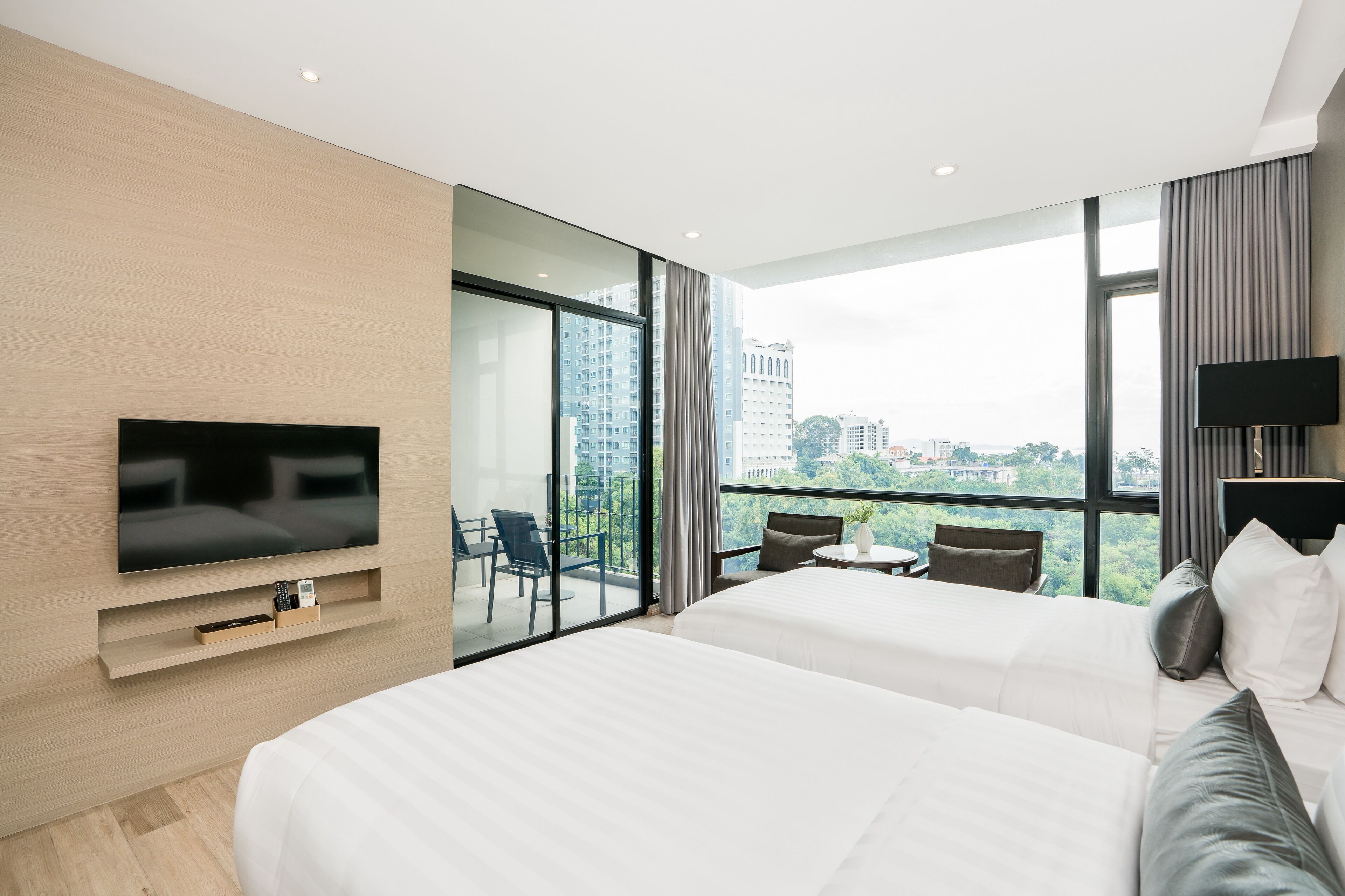 A modern hotel room with a large bed, a TV, a balcony with a view of the city, and a small table with chairs.