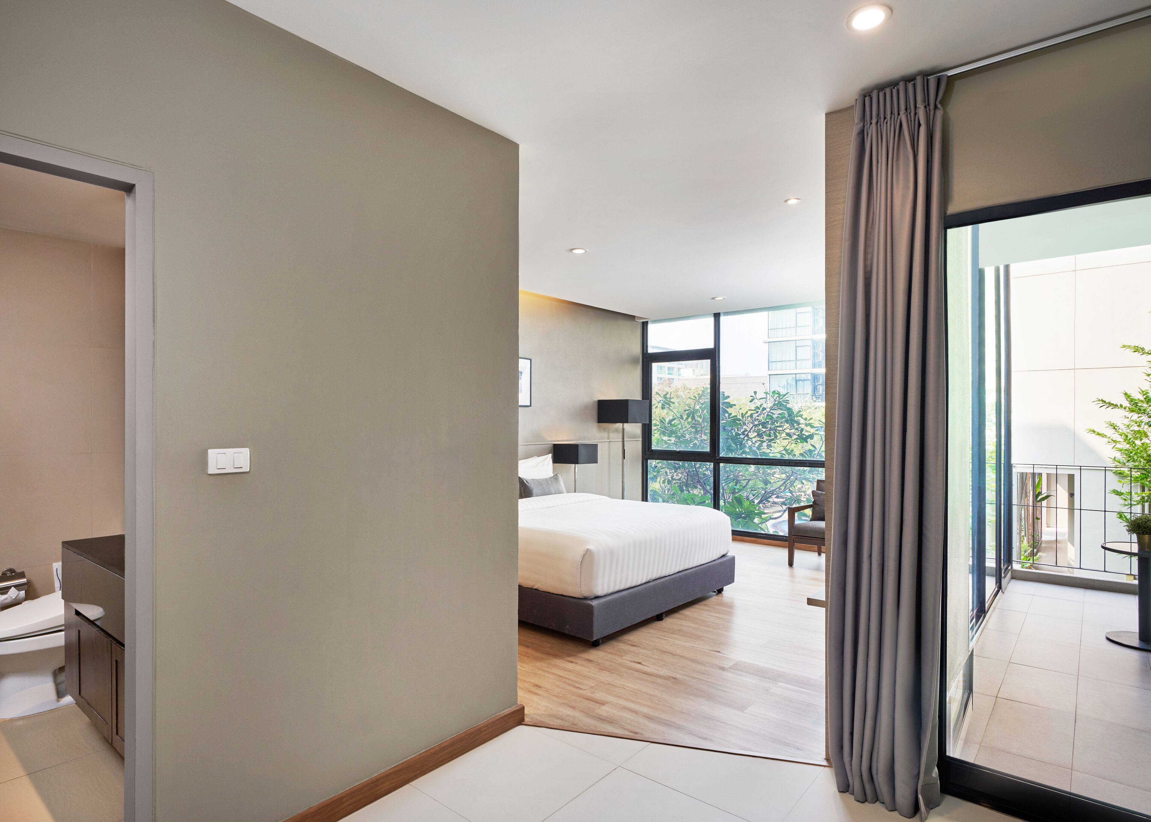 A modern hotel room with a large bed, a bedside table, a chair, and a view of the outdoors through a large window.