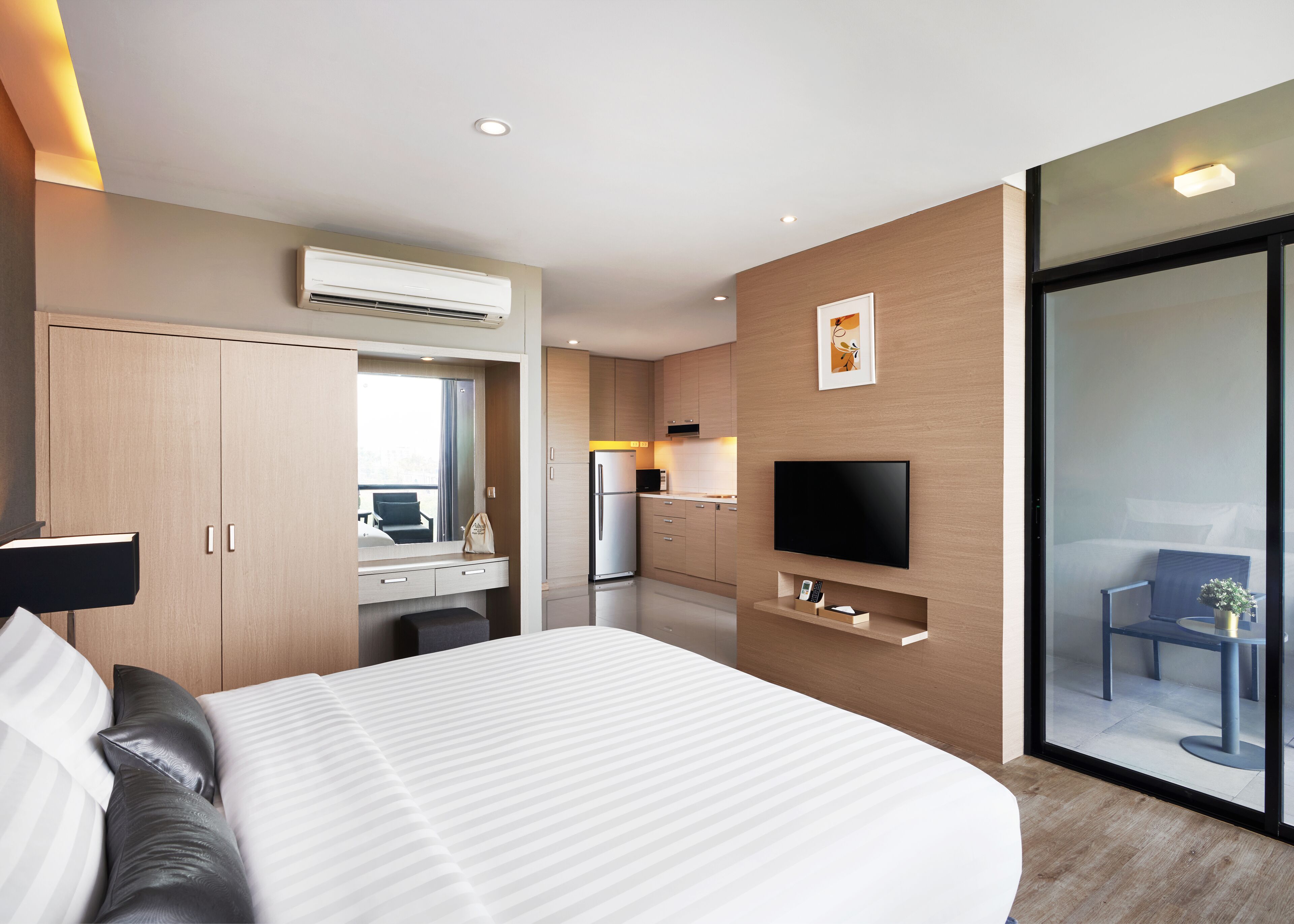 Una habitación de hotel moderna con una cama grande, un escritorio con computadora, televisor de pantalla plana, zona de cocina y puerta de vidrio corredera al balcón.