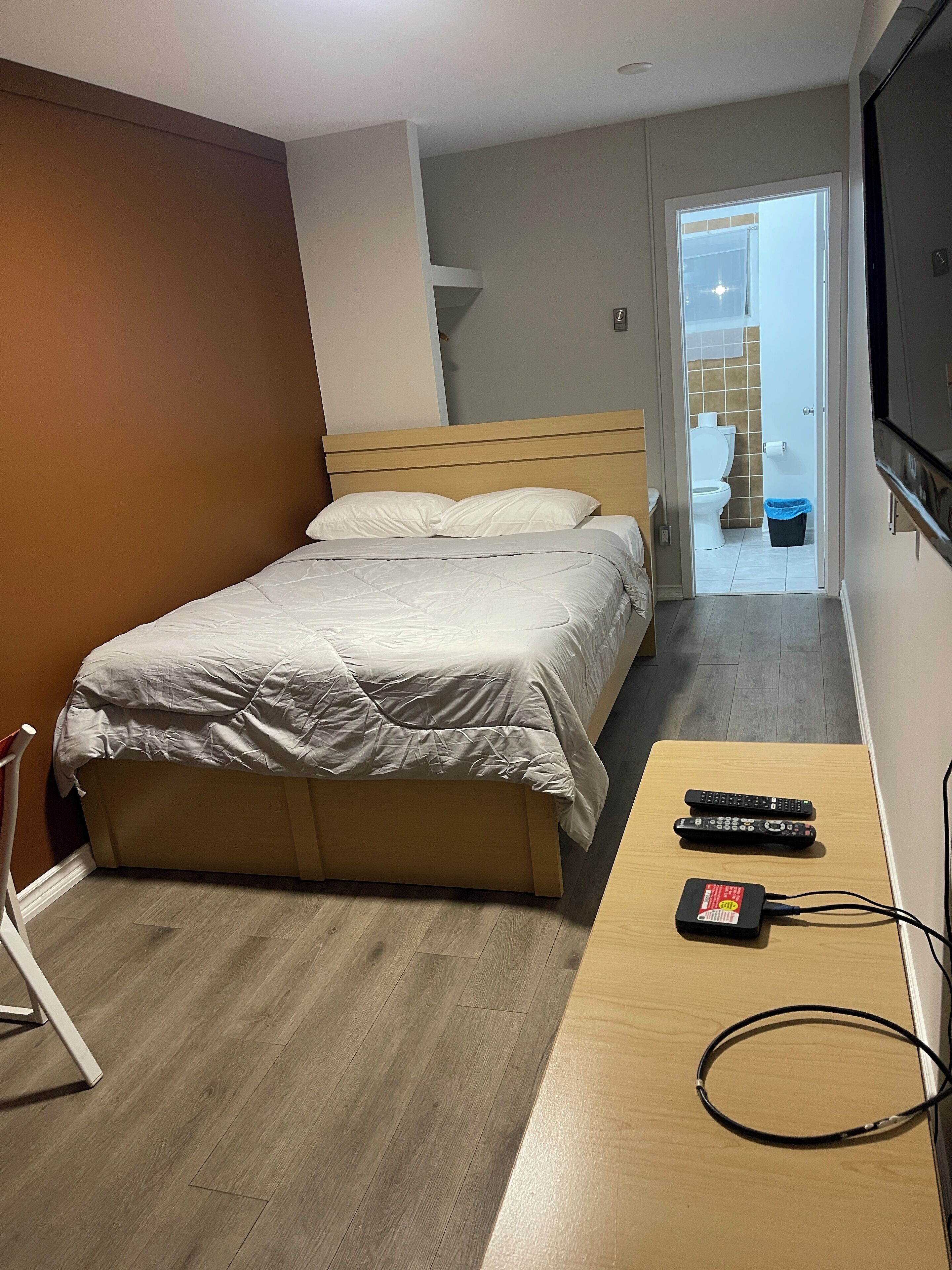 A bedroom with a bed, a wooden bedside table with a power strip and remote controls, a TV mounted on the wall, and a bathroom visible through an open door.