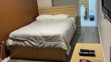 A bedroom with a bed, a wooden bedside table with a power strip and remote controls, a TV mounted on the wall, and a bathroom visible through an open door.