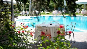 Un área de comedor junto a la piscina con mesas y sillas redondas, rodeada de una piscina y follaje.