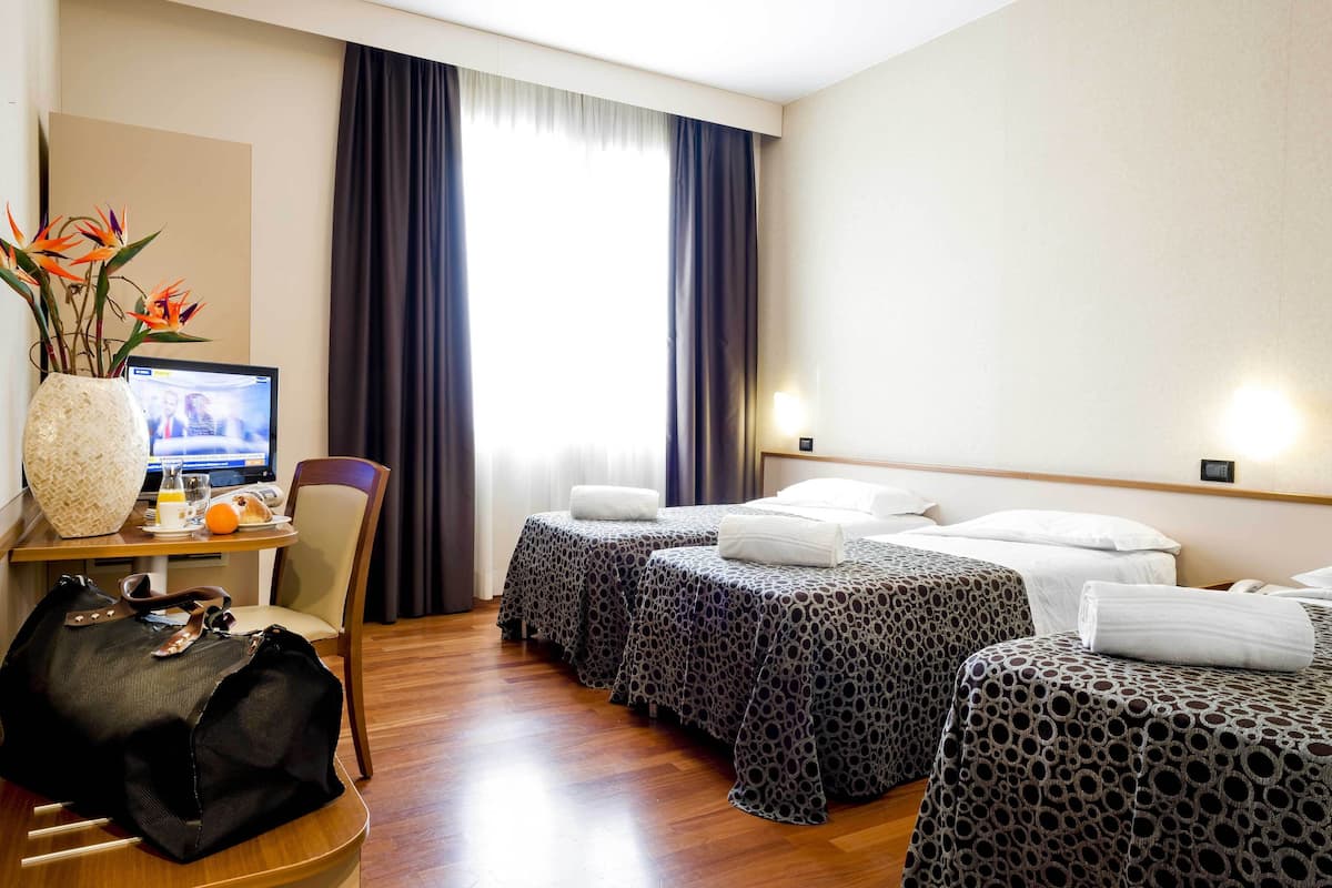 A hotel room with two beds, a desk with a computer, a chair, a large vase with flowers, and a window with curtains.