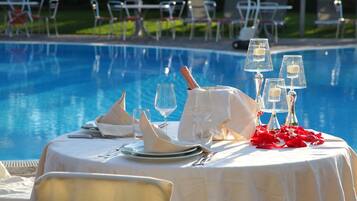 Mesa lista para una comida junto a la piscina, con una botella de champán, copas y pétalos de rosa roja.