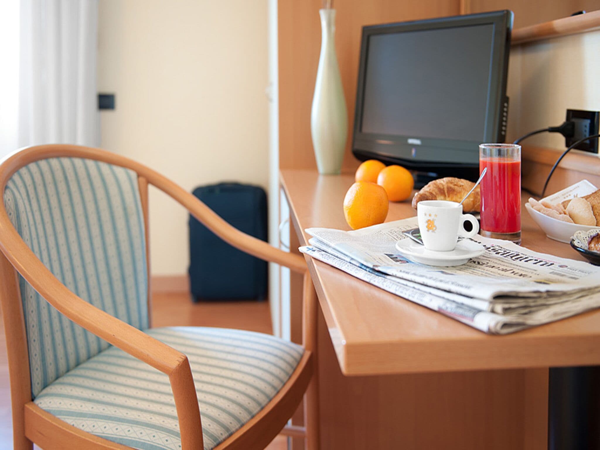 Une chambre d’hôtel avec une chaise en bois, une petite table sur laquelle sont disposés des articles pour le petit-déjeuner, une télévision et une valise.