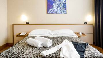 Una camera d'albergo con una coperta a motivi, asciugamani piegati con cura, un telefono sul comodino e un quadro appeso al muro.