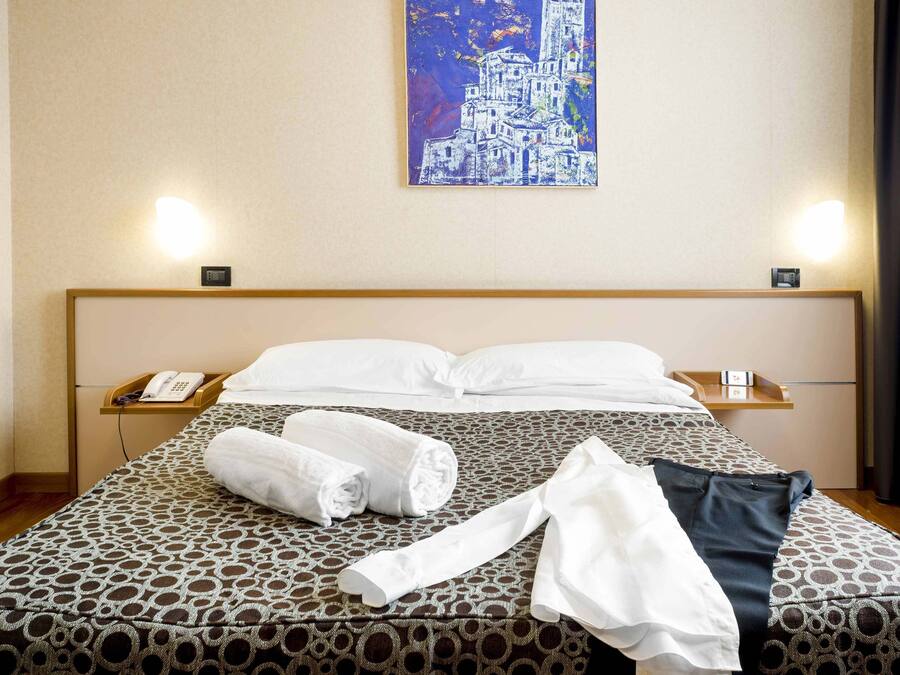 Een hotelkamer met een bed met een patroon, netjes opgevouwen handdoeken, een telefoon op het nachtkastje en een schilderij aan de muur.