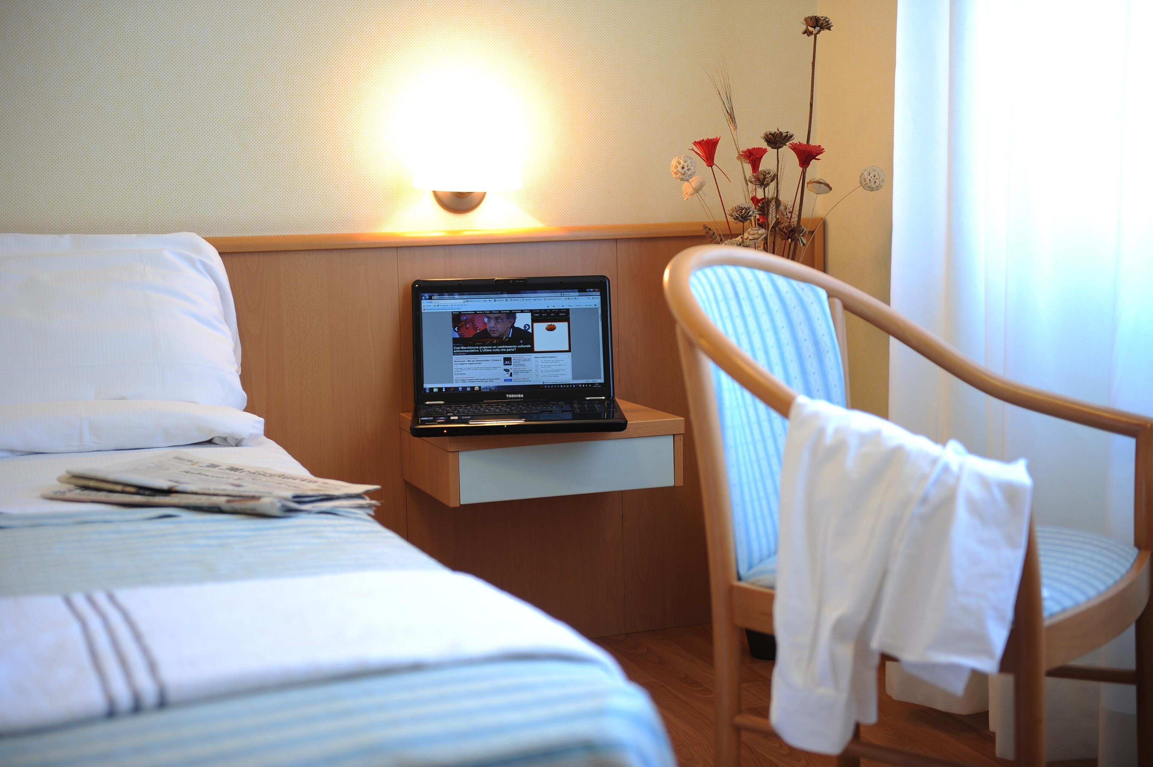 A hotel room with a bed, a laptop on a bedside table, a chair, and a vase with flowers.